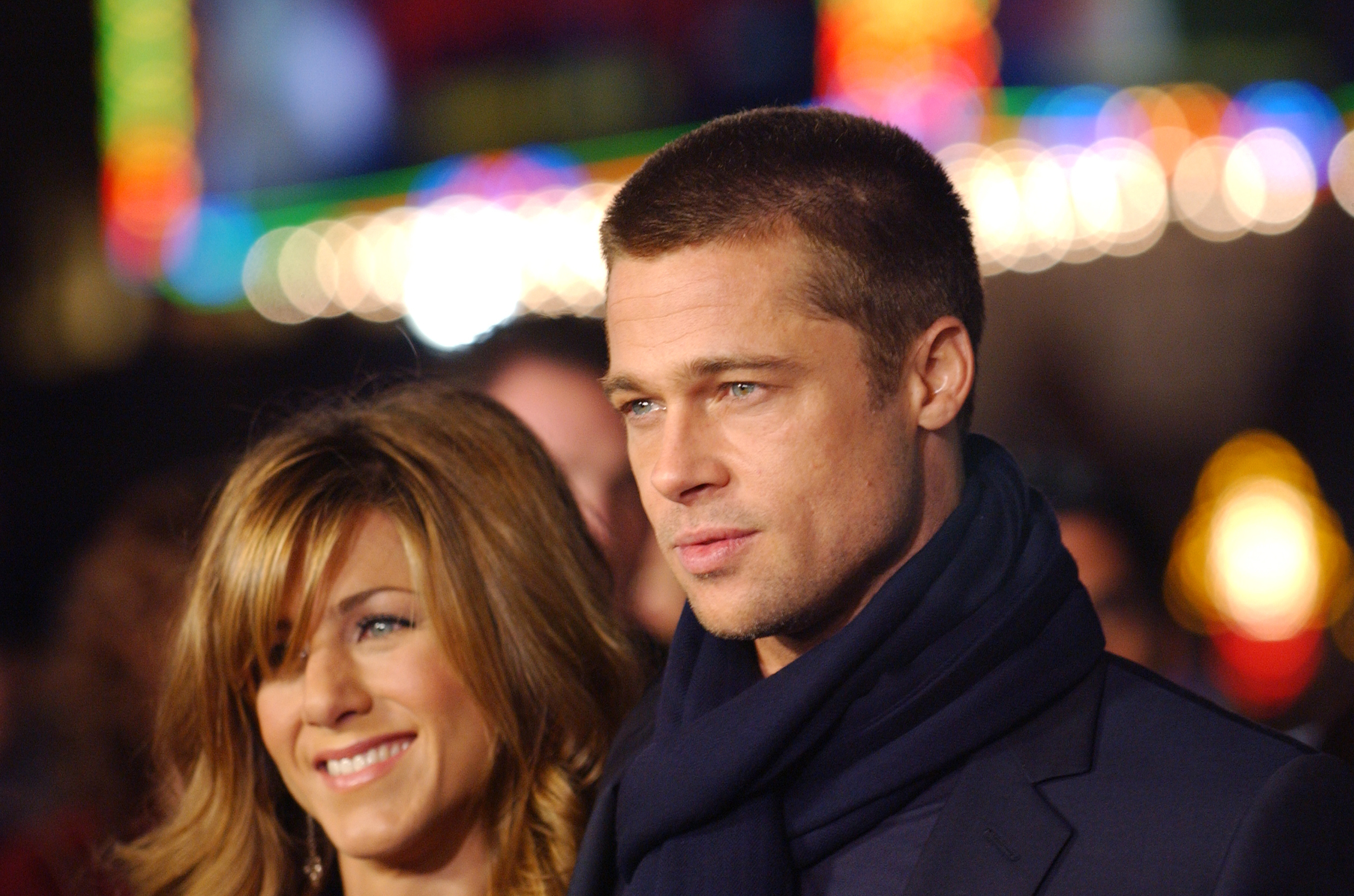 Jennifer Aniston smiles beside Brad Pitt during the "Along Came Polly" Los Angeles premiere, with colorful lights and blurred crowds glowing behind the couple.