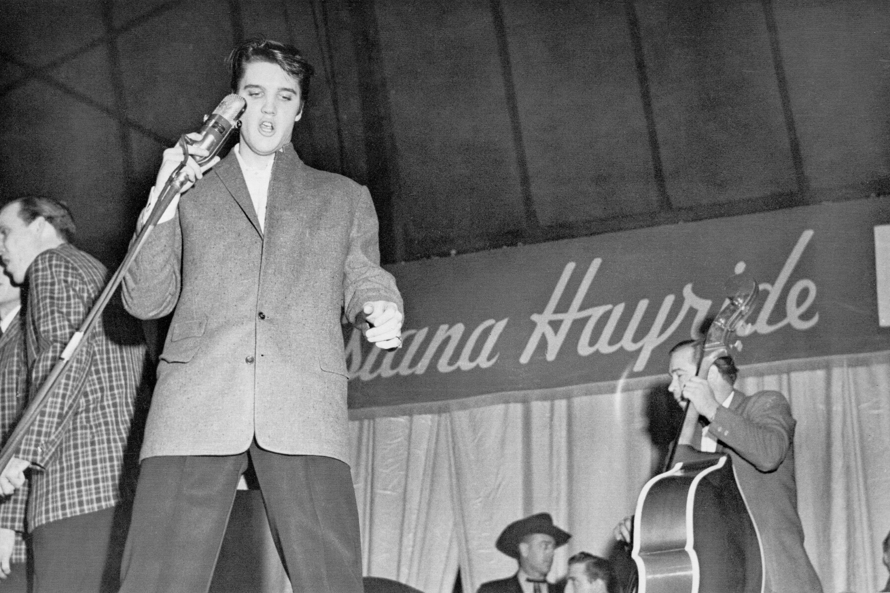 Rock and roll singer Elvis Presley sings into a vintage microphone alongside bass player Bill Black while performing on tour with the "Louisiana Hayride" show in 1954, wearing a tailored single-breasted tweed suit jacket over dark trousers and a crisp white collared shirt. | Source: Getty Images