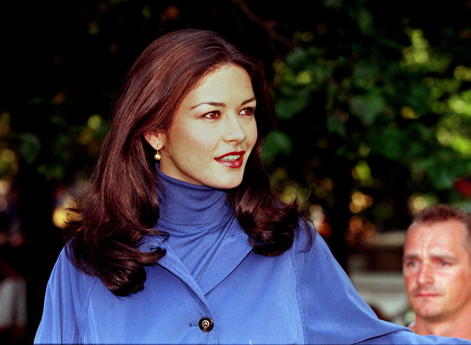 Catherine Zeta-Jones pauses on set in South East London during the filming of "Entrapment" on August 11, 1998. She wears an all-blue ensemble consisting of a high-neck turtleneck layered beneath a buttoned trench coat, accessorized with pearl drop earrings, a berry lip, and styled blowout waves.