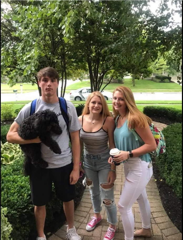 On a leafy front walkway, Jack Pugh stands with two friends and a small black dog in his arms. | Source: Facebook/KennaLynMcClelland