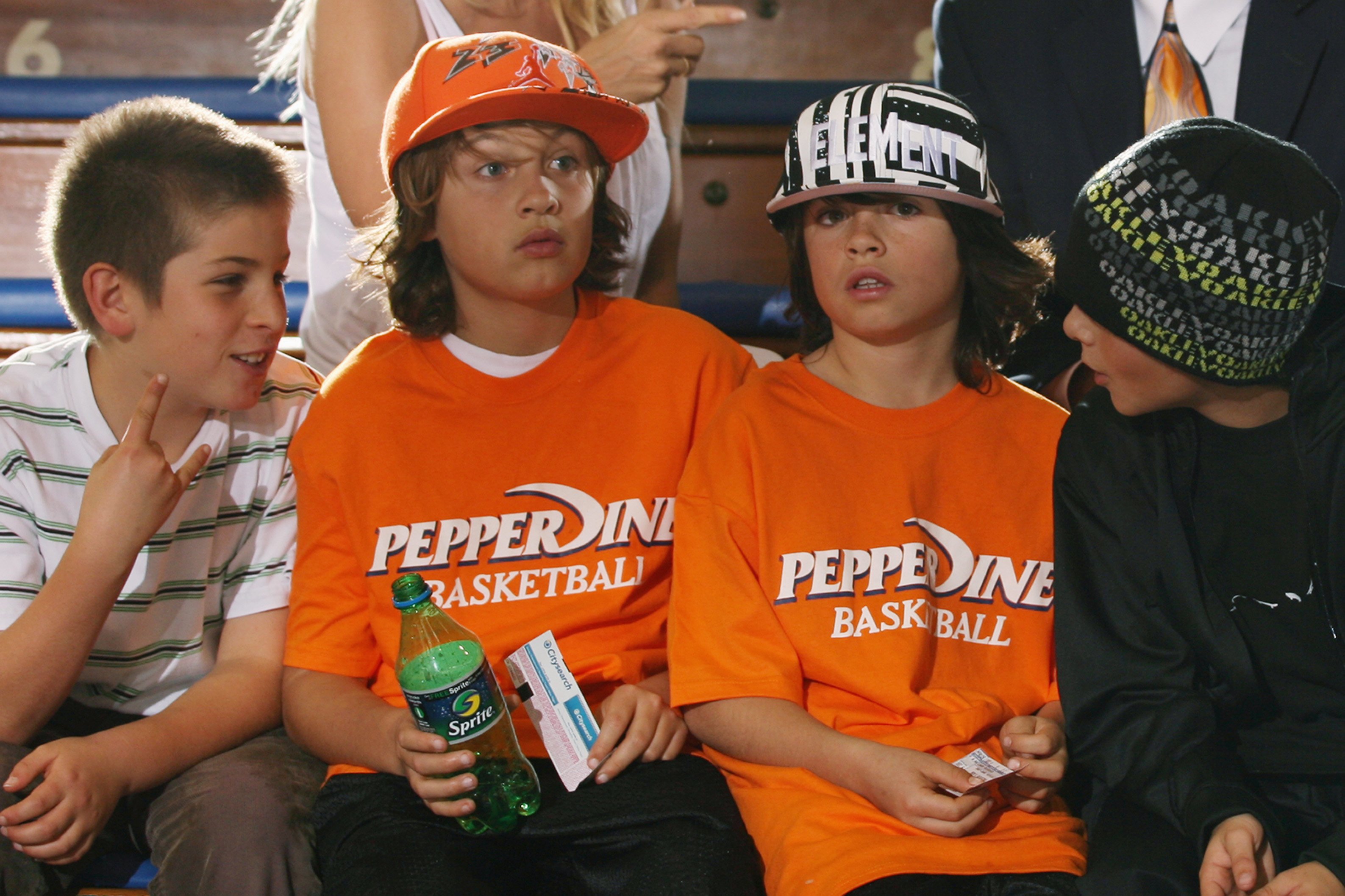 Brandon (wearing an orange cap) and Dylan (in a striped cap) sit with friends before the basketball game between the Loyola Marymount Lions and the Pepperdine Waves on January 26, 2008, at Firestone Fieldhouse in Malibu, California. Both boys wear matching orange Pepperdine Basketball T-shirts as they watch from the stands.