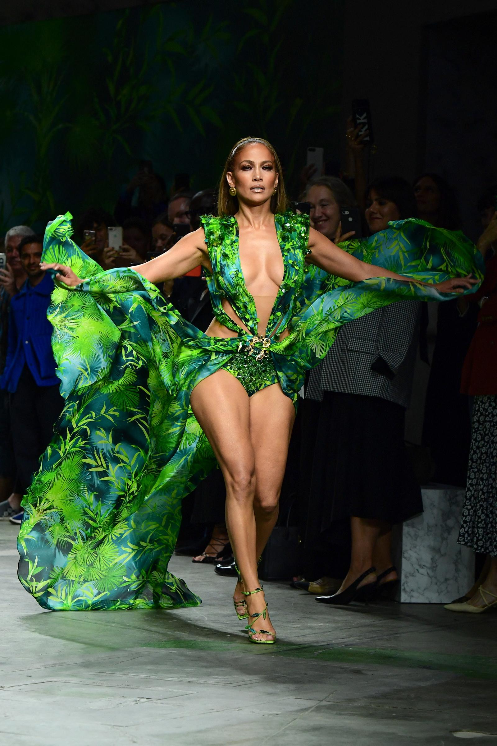 Jennifer Lopez commands the runway in a dramatic tropical-print Versace look, stretching both arms outward as flowing green fabric billows behind her. The plunging outfit, matching heels, and powerful stride transform the fashion show appearance into a theatrical moment.
