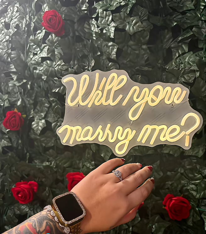 Pumpkin shows off her sparkling engagement ring beneath a glowing “Will you marry me?” sign surrounded by red roses and green foliage. The close-up highlights the ring, her red manicure, and her smartwatch, offering a closer look at the jewelry from Darrin’s proposal. | Source: TikTok/Mama P