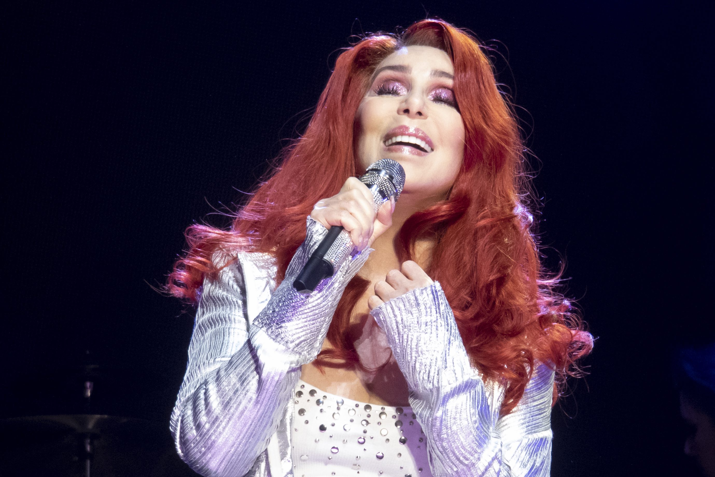 Cher belted into a microphone on stage during her Here We Go Again Tour stop at the SSE Hydro Arena in Glasgow, United Kingdom, on October 28, 2019. The pop icon showcased a dramatic, fiery red-orange wig styled with voluminous, swept-back root lift and soft, cascading waves that draped across her shoulders, complementing her shimmering pink eyeshadow and metallic silver outfit. | Source: Getty Images