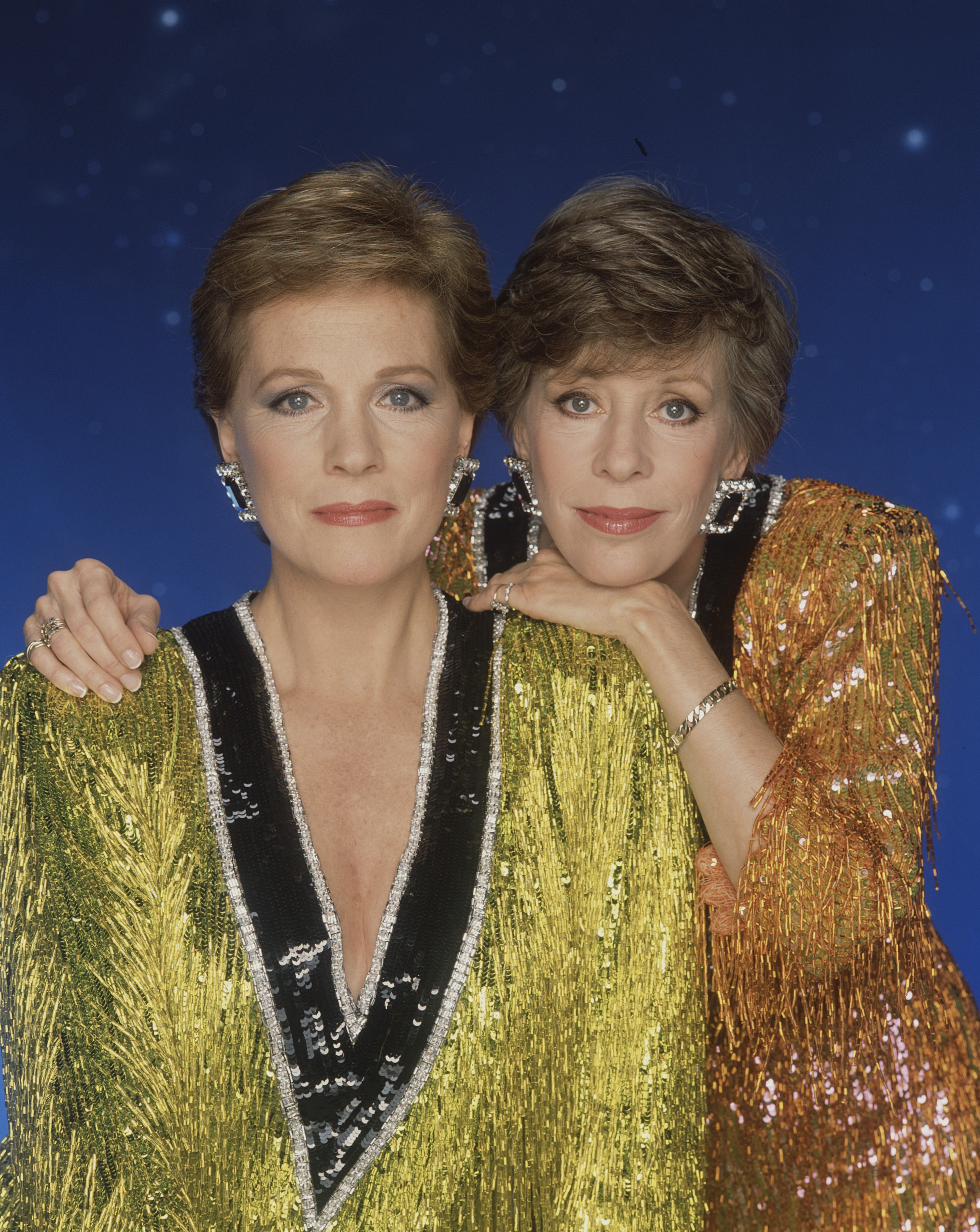 Julie Andrews poses alongside longtime friend and collaborator Carol Burnett in a promotional portrait for the 1989 ABC television special "Julie & Carol: Together Again," recorded at the Pantages Theatre in Hollywood. The dynamic duo dazzle against a starry blue backdrop in shimmering sequined ensembles, with Andrews wearing a striking lime-gold tinsel gown featuring a plunging black-and-silver sequin V-neck trim and statement crystal earrings while Burnett rests affectionately against her shoulder.