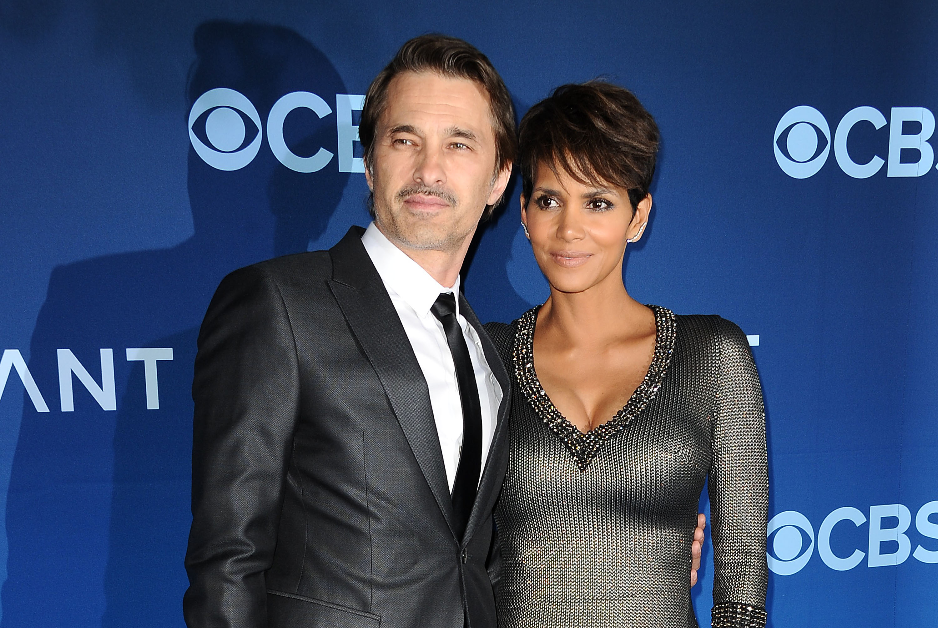 Actor Olivier Martinez and actress Halle Berry posed together on the red carpet at the premiere of "Extant" held at the California Science Center on June 16, 2014, in Los Angeles, California. Martinez looked sharp in a dark charcoal tailored suit, crisp white dress shirt, and slim black tie, while Berry complemented him in a metallic textured gunmetal knit dress featuring a sparkling beaded neckline and her signature cropped pixie cut.