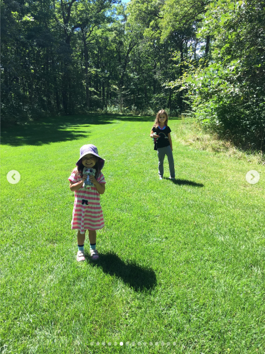 Mabel and Fiona enjoy a sunny day outdoors in a wide grassy field surrounded by trees, with Fiona in a pink-and-white striped dress, purple sun hat and sandals and Mabel standing behind her in a dark top and gray pants. The sunlight casts long shadows across the grass, capturing a peaceful childhood moment. | Source: Instagram/mabellowe_