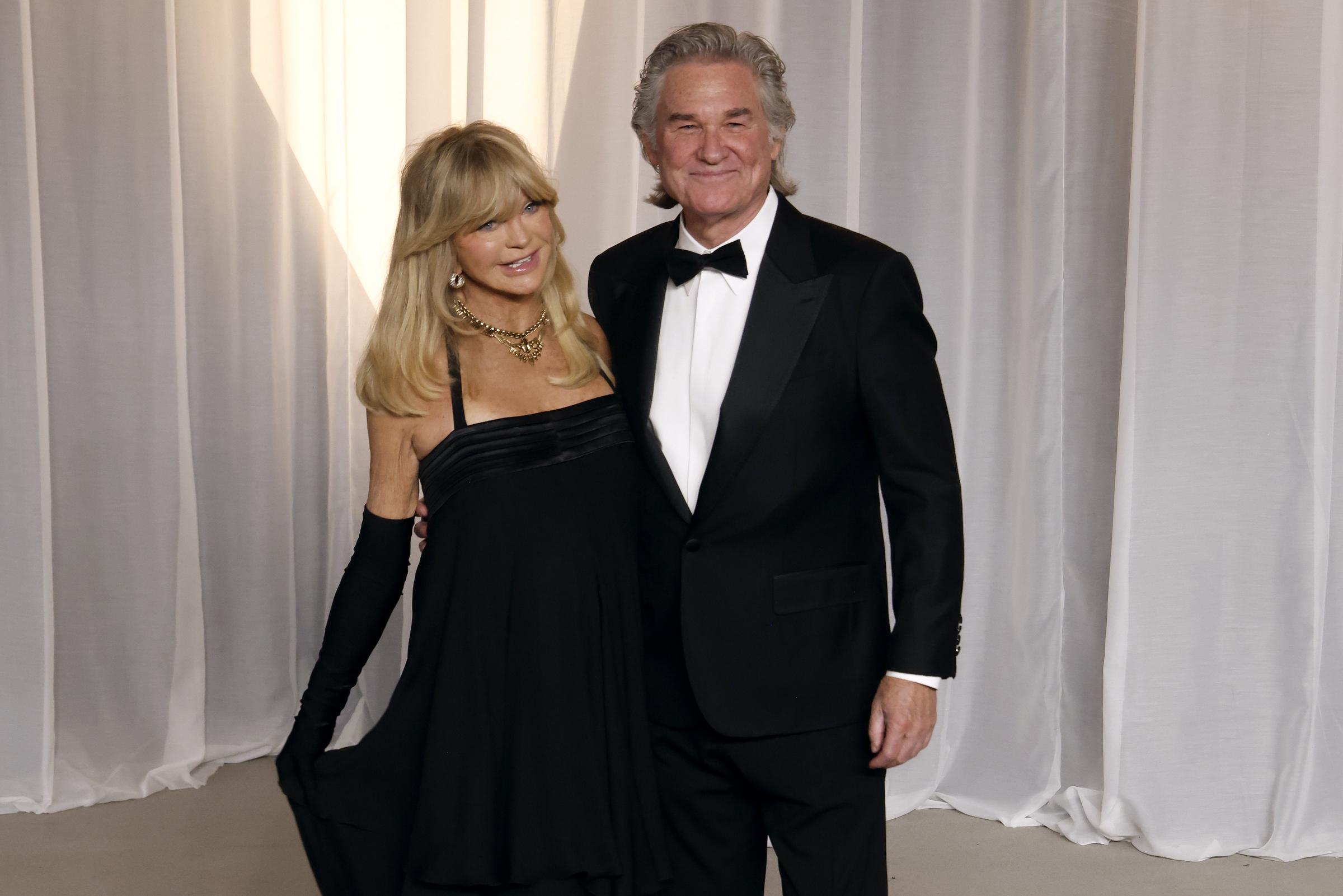 Goldie Hawn and Kurt Russell make an effortlessly chic pair in coordinating formal black attire. Hawn charms in an empire-waist black cocktail dress styled with matching opera-length gloves, statement gold jewelry, and her signature voluminous blonde bangs. Russell complements her classic style in a sharp black tuxedo featuring peaked satin lapels, a crisp white dress shirt, and a traditional black bow tie.