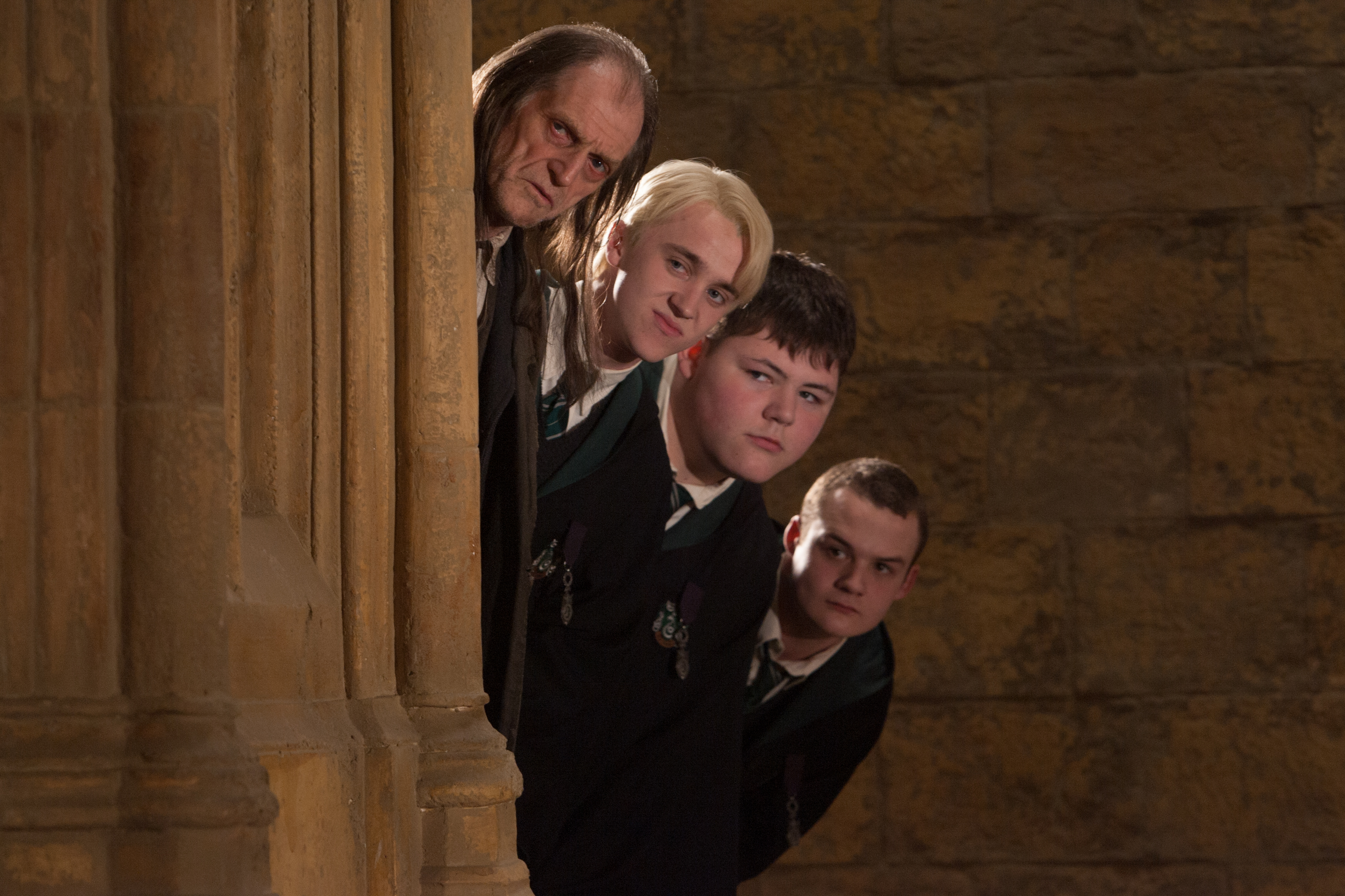 David Bradley, Tom Felton, Jamie Waylett, and Josh Herdman peer around a stone pillar like a mischievous Slytherin totem pole in motion. Their expressions range from sly to suspicious, each one perfectly in character against the cold Hogwarts walls. It's a moment of on-set camaraderie wrapped in classic villainous curiosity from "Order of the Phoenix."