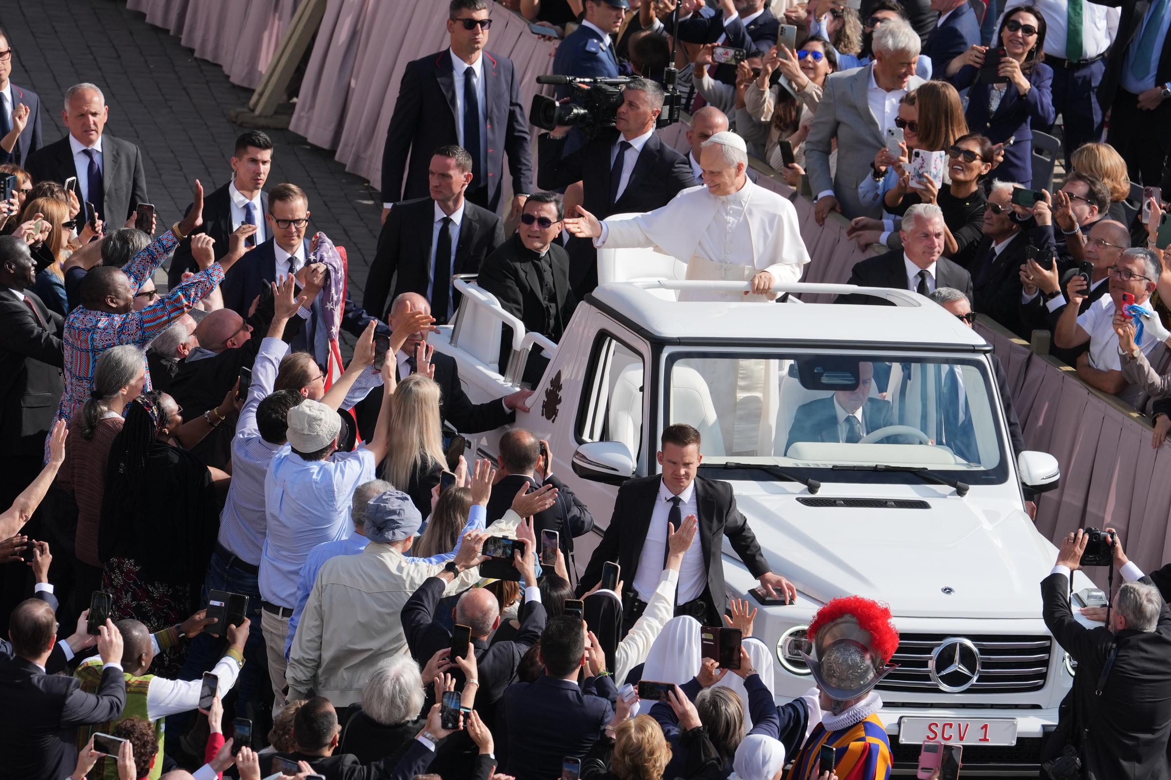 Pope Leo XIV waves to an enthusiastic crowd as he rides through St. Peter's Square in an open Popemobile on May 18, 2025, during his inauguration mass, as attendees cheer and take photos of the newly elected pontiff.