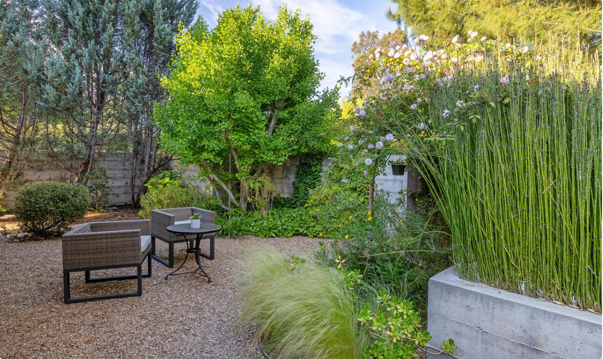 A secluded garden patio features woven outdoor chairs, a small table, flowering plants, and tall bamboo that adds privacy to the landscaped space. | Source: Redfin
