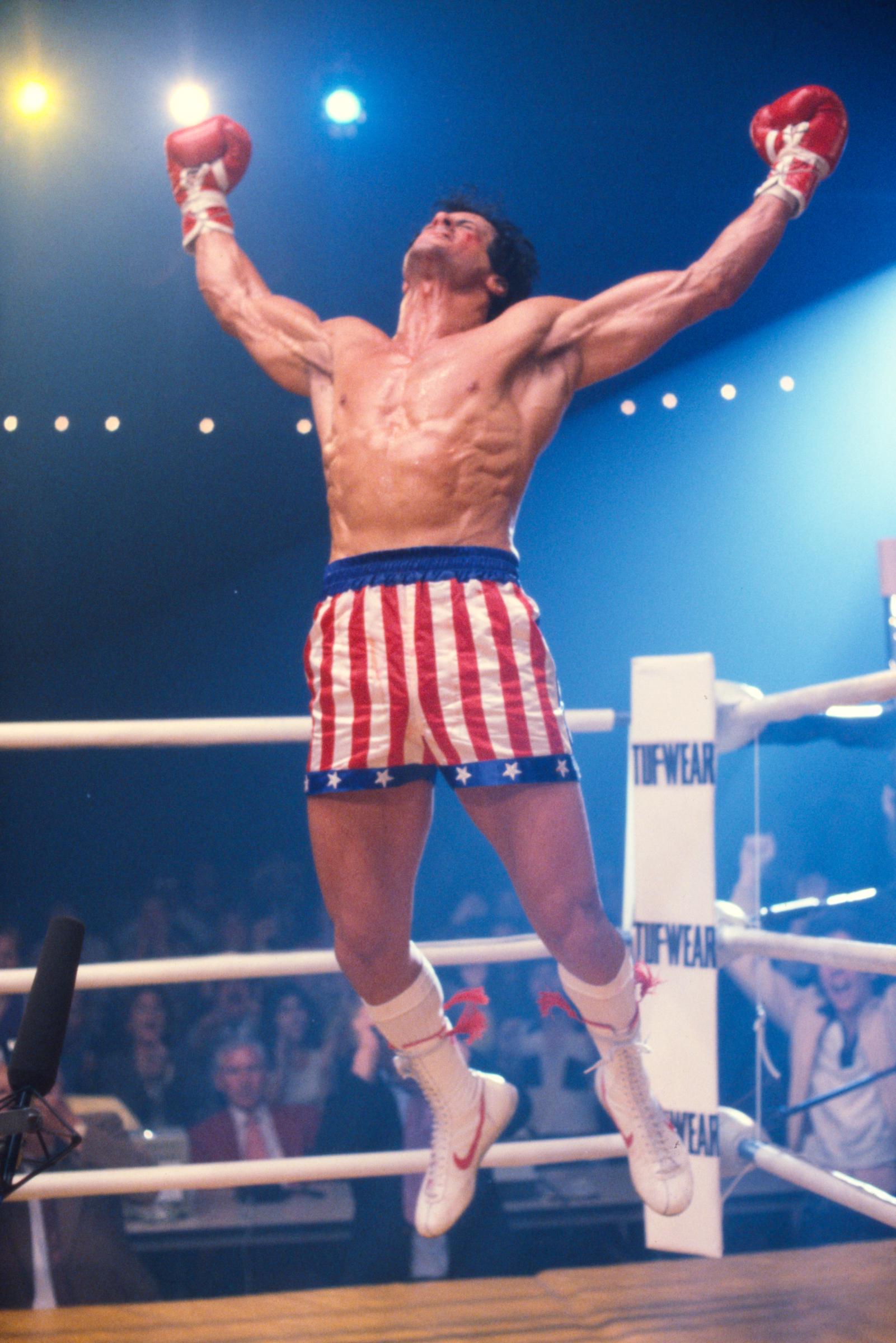 Sylvester Stallone, as Rocky Balboa, leaps into the air with his gloves raised in triumph on the boxing set of "Rocky III" in Los Angeles, California, in May 1982. Wearing Rocky's signature red, white, and blue trunks, Stallone displays the sharply defined physique he achieved through his demanding workout routine and restrictive diet.