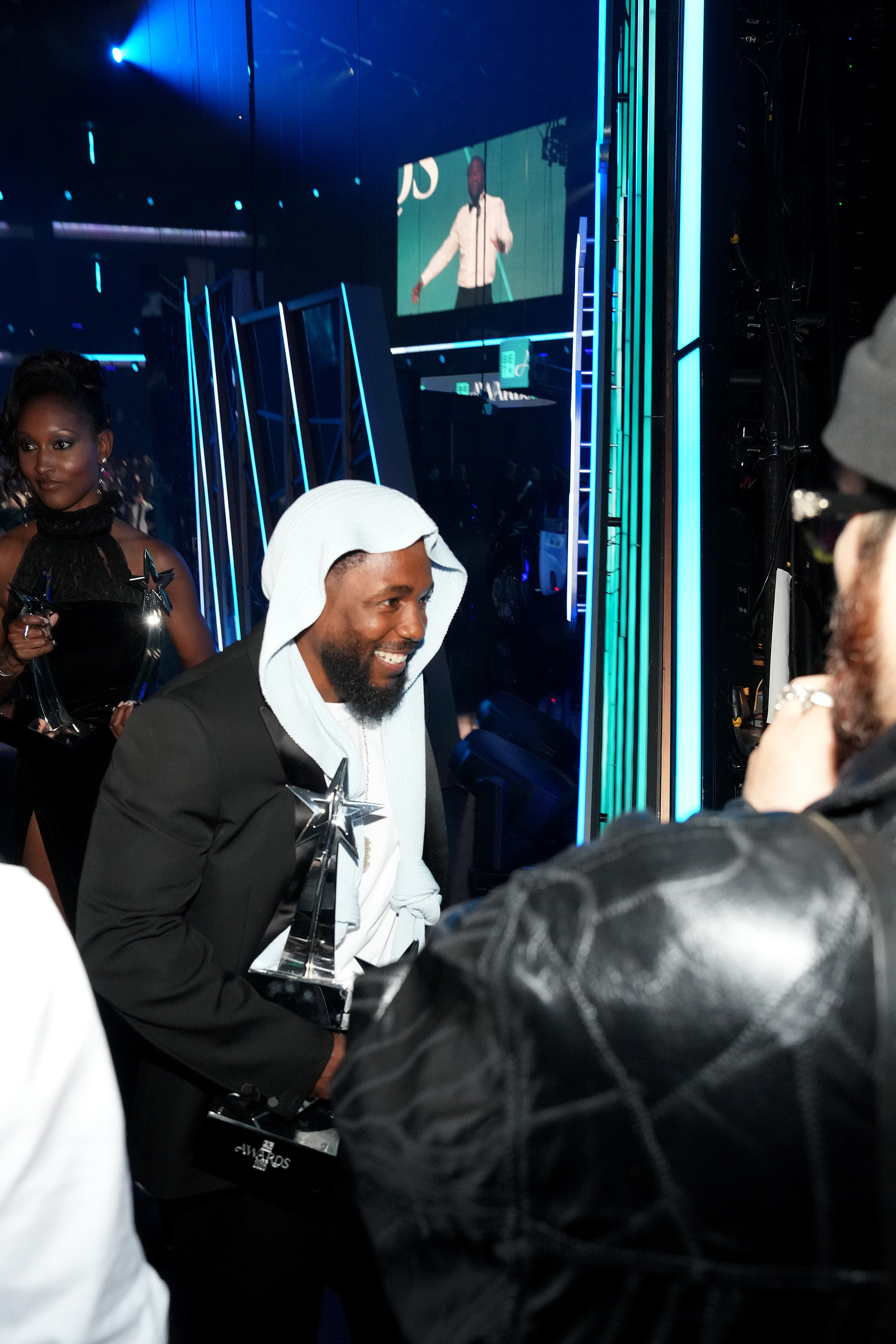 Backstage at the Peacock Theater, Kendrick Lamar is all smiles as he greets well-wishers and clutches multiple awards following his wins at the 2025 BET Awards. His layered look included a knit headpiece tied at the front.