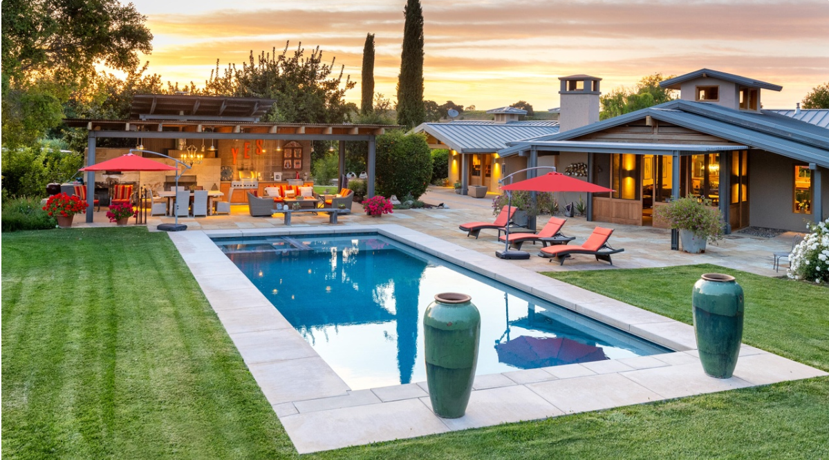 The property features a swimming pool surrounded by a stone patio, sun loungers, and a covered outdoor kitchen and dining area. | Source: Redfin