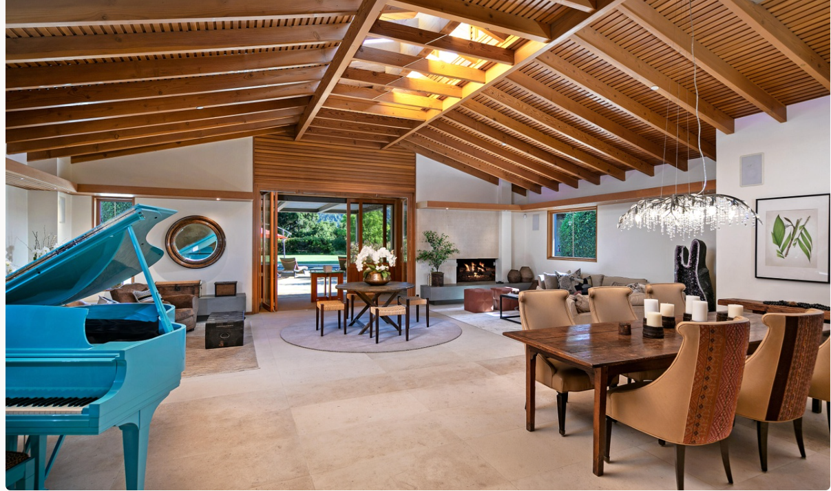 The home's open-plan living and dining room features a vaulted wooden ceiling, a turquoise grand piano, a fireplace, and wide glass doors overlooking the garden. | Source: Redfin