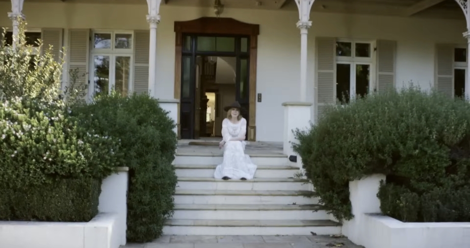 Framed by manicured hedges and classic white pillars, the front steps of the farmhouse offer a stately yet inviting entry to the property’s grand colonial architecture. From a video dated July 20, 2015. | Source: YouTube/Vogue