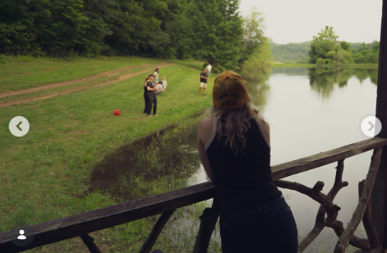 A person leans against a wooden railing overlooking a pond bordered by grass and trees. The image adds a different perspective to Wynonna's outdoor collection, shifting from the house and porch to the surrounding landscape. | Source: Instagram/wynonnajudd
