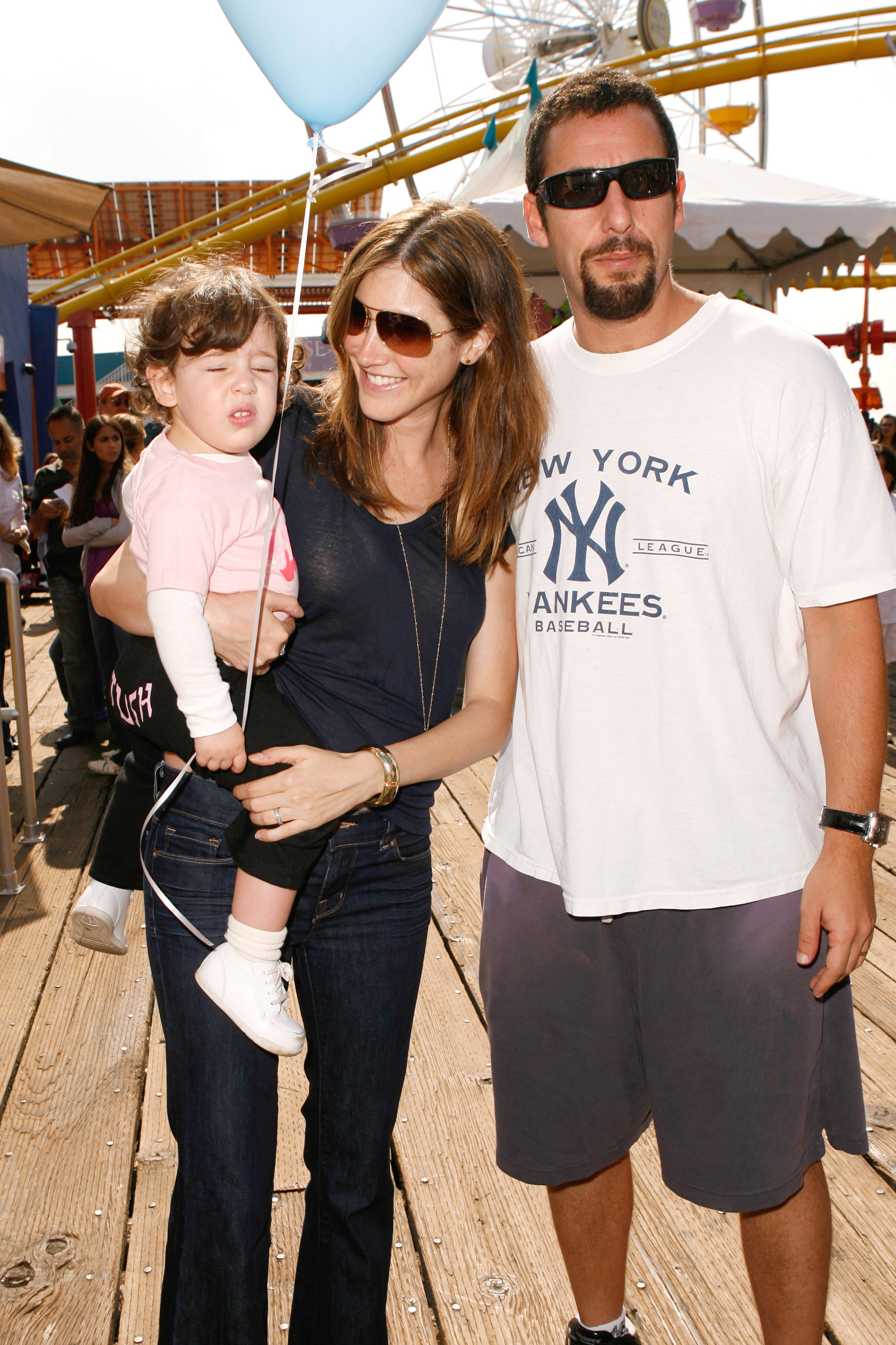 Adam Sandler stands beside Jackie Sandler, who holds baby Sadie Madison Sandler, as they enjoy a sunny day out together. Dressed casually, the family shares a relaxed, candid moment — blending everyday warmth with a glimpse into their off-screen life.