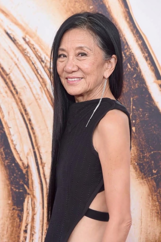 Embracing a conventional aging journey, this AI rendering pictures Vera Wang with delicate crow's feet, laugh lines, and fine silver threads woven into her sleek side-swept locks. Posing in front of a swirling neutral backdrop reminiscent of high-fashion award arrivals, the simulated designer shines in a sleeveless black dress featuring dramatic open-side architecture and an elongated sparkling crystal drop earring. | Source: Gemini