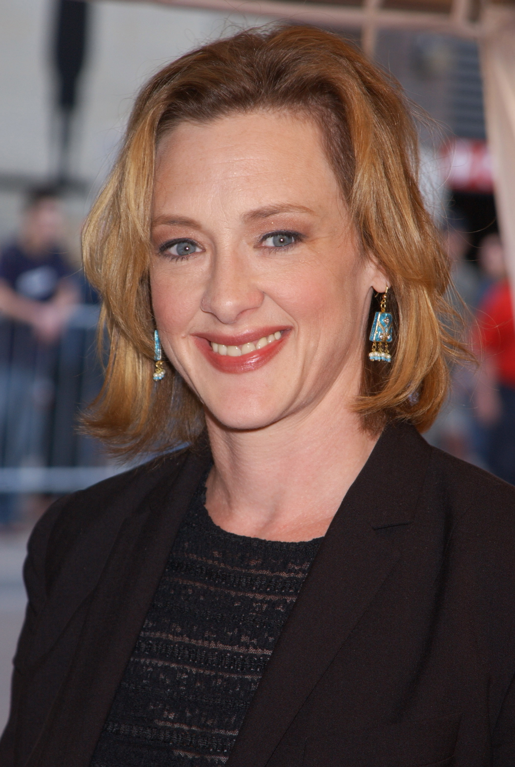Wearing a black blazer over a textured top, Joan Cusack smiles brightly while posing for photographers. Colorful earrings bring a playful contrast to the otherwise understated outfit.