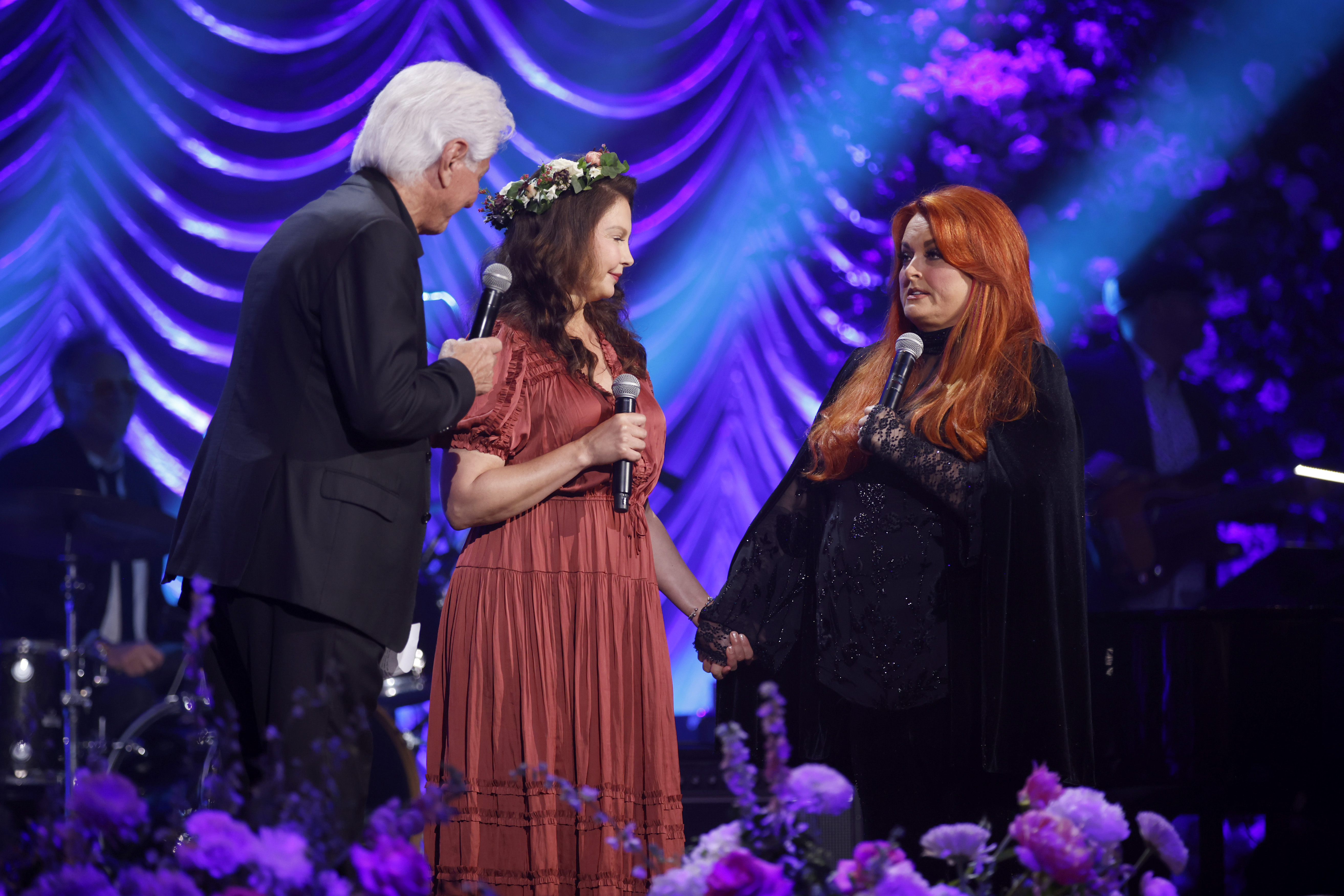 Ashley and Wynonna Judd hold hands during their mother's memorial at Nashville's Ryman Auditorium on May 15, 2022. Their stepfather, Larry Strickland, joins them onstage. | Source: Getty Images
