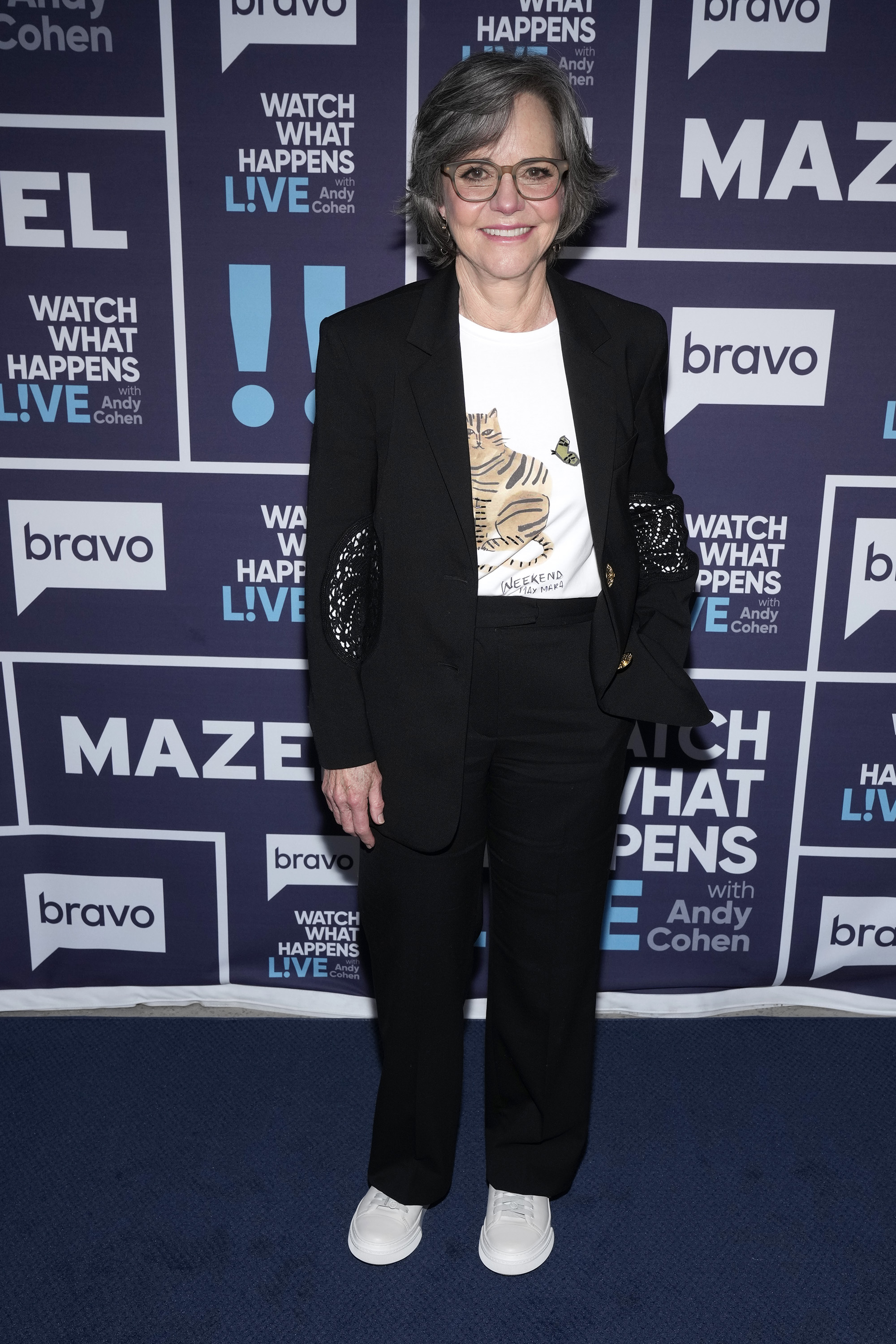 Sally Field on "Watch What Happens Live with Andy Cohen," looking effortlessly cool in a semi-formal black suit paired with sneakers and a white printed T-shirt, smiling gently at the camera.