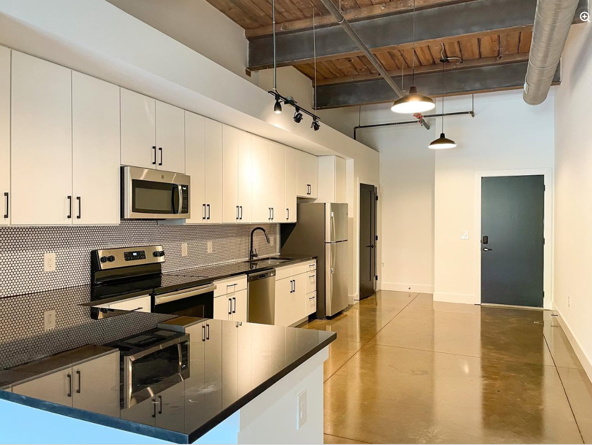 The open-plan kitchen pairs white cabinetry and stainless-steel appliances with a black-and-white backsplash, while exposed beams and ductwork overhead preserve the loft's industrial character. | Source: Facebook/Judson Mills Loft