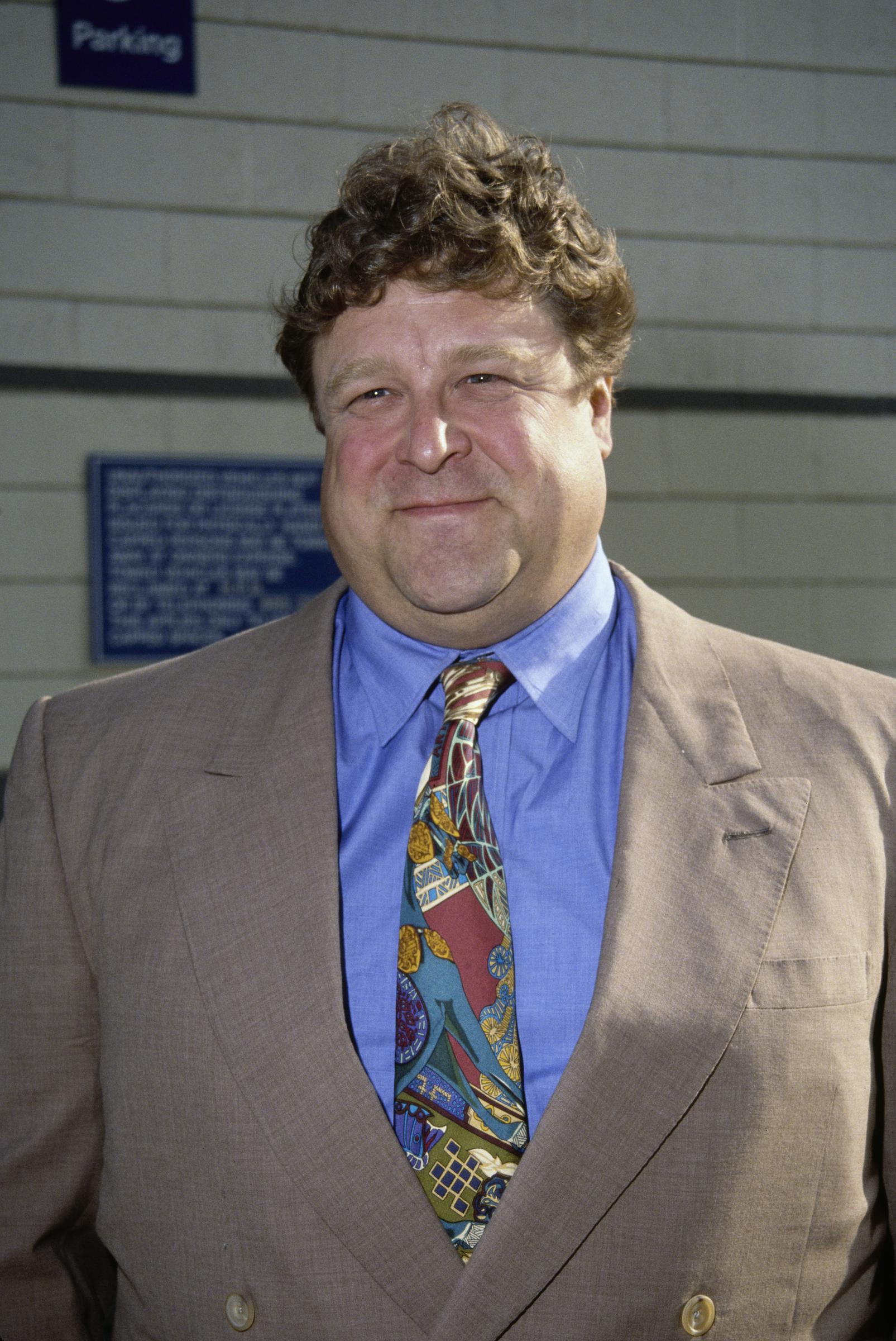 John Goodman appears in a brown suit, blue shirt and colorful patterned tie, offering an early look at the actor long before his major weight loss. The portrait captures the fuller appearance longtime viewers came to associate with him during that period.