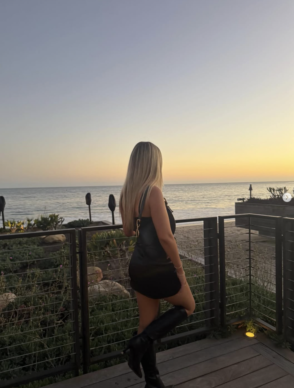 Finley Lockwood takes in the sunset from an oceanfront terrace, pictured from behind in a sleek black sleeveless mini dress and knee-high black leather boots, with her long blonde hair cascading down her back. | Source: Instagram/finleyl.ockwood