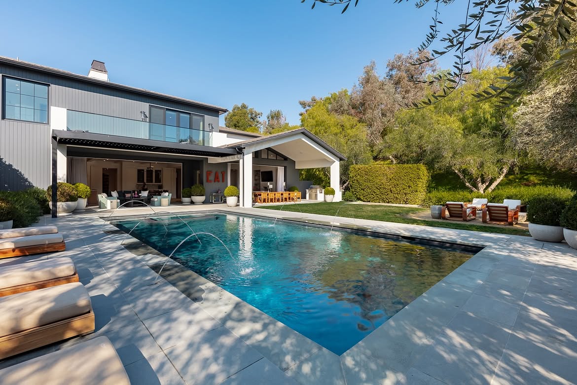 The rear view shows how the pool sits between the house and its landscaped grounds. Covered living and dining spaces open toward the water, creating a continuous entertaining area that stretches from the interior into the backyard. | Source: Instagram/tomer.fridman
