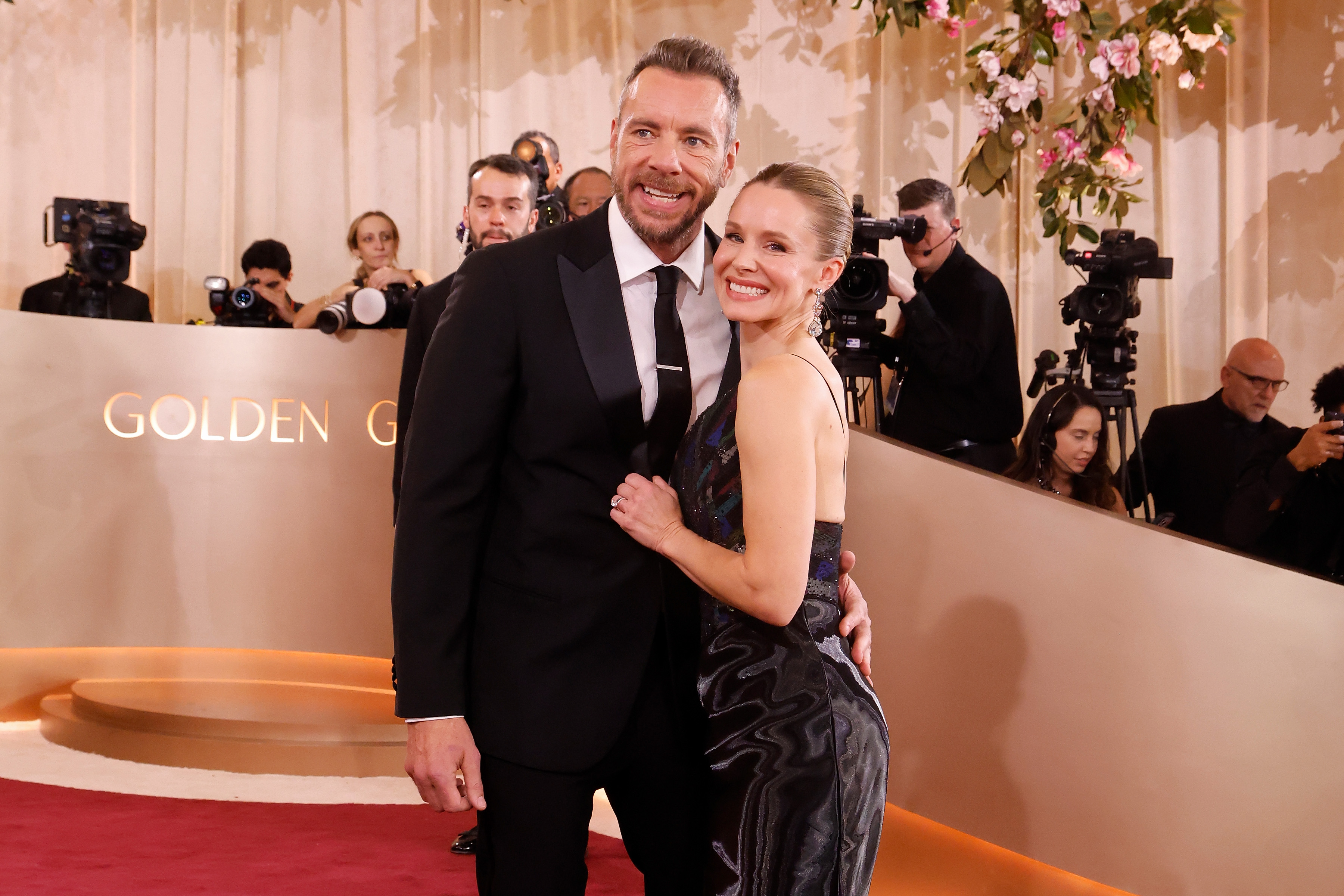 Dax Shepard stands with a relaxed smile, looking off-camera, as Kristen Bell beams with joy beside him. The couple shares a sweet moment together on the Golden Globes red carpet.