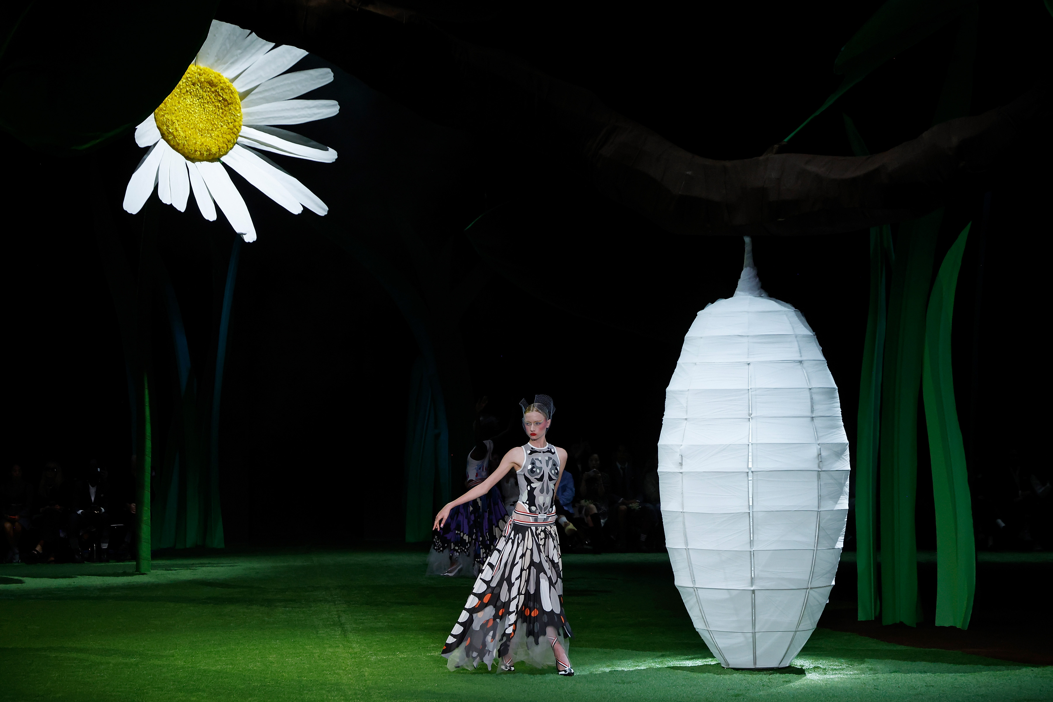 Kai Schreiber glided across a whimsical grassy set under a suspended oversized daisy and lantern cocoon during the Thom Browne September 2026 fashion show at The Shed on September 15, 2026, in New York City. Making an ethereal runway appearance during New York Fashion Week, the model presented a graphic grey-and-black bodysuit paired with a flowing, layered butterfly-wing printed skirt edged in sheer tulle. | Source: Getty Images