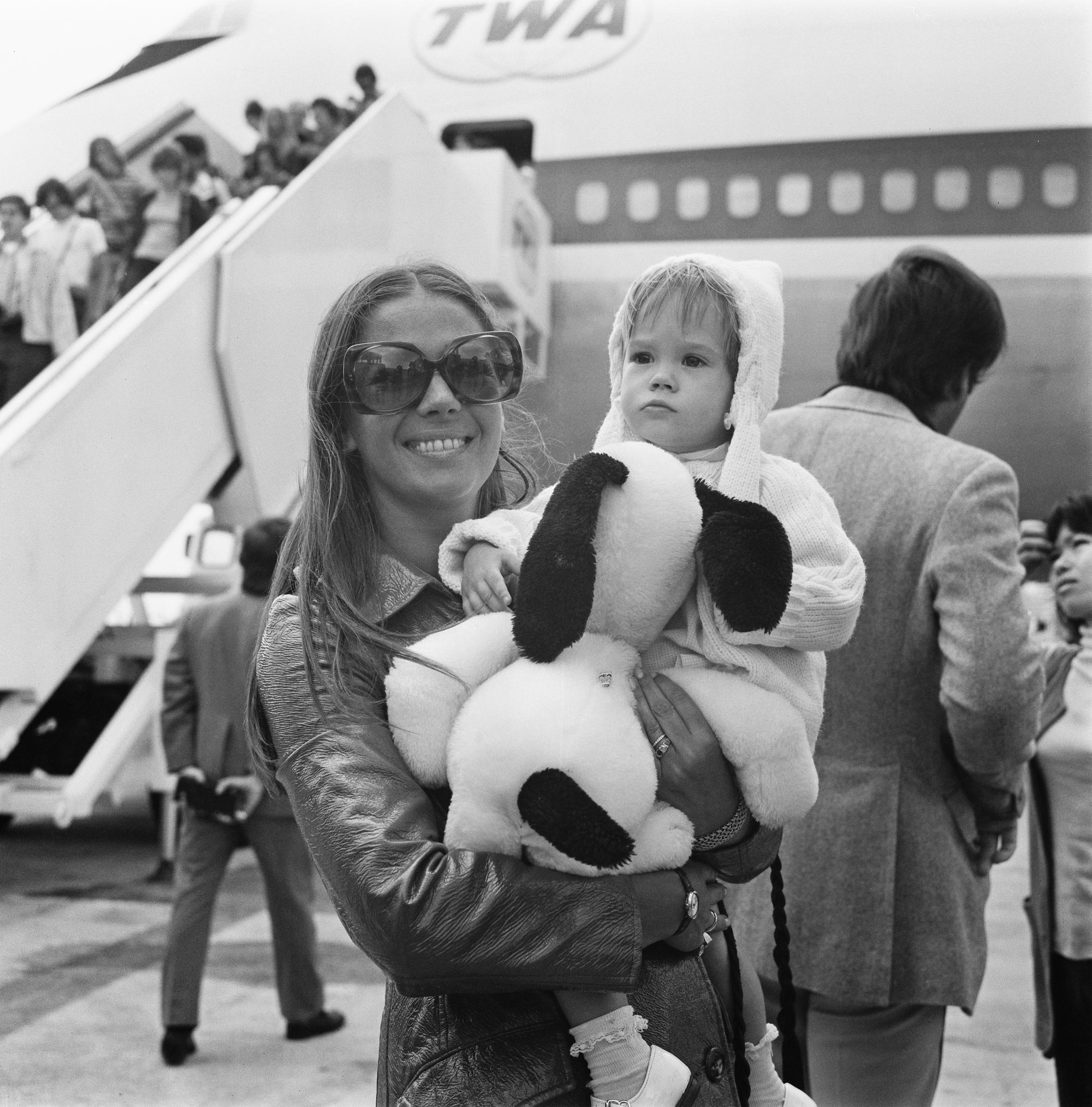 Natalie Wood stands at Heathrow Airport holding her young daughter Natasha Gregson Wagner in her arms, on August 4, 1972. The black-and-white image shows Natalie wearing a leather jacket and oversized sunglasses as she smiles at the camera. Natasha, bundled in a hooded sweater, clutches a plush Snoopy toy and looks off to the side.