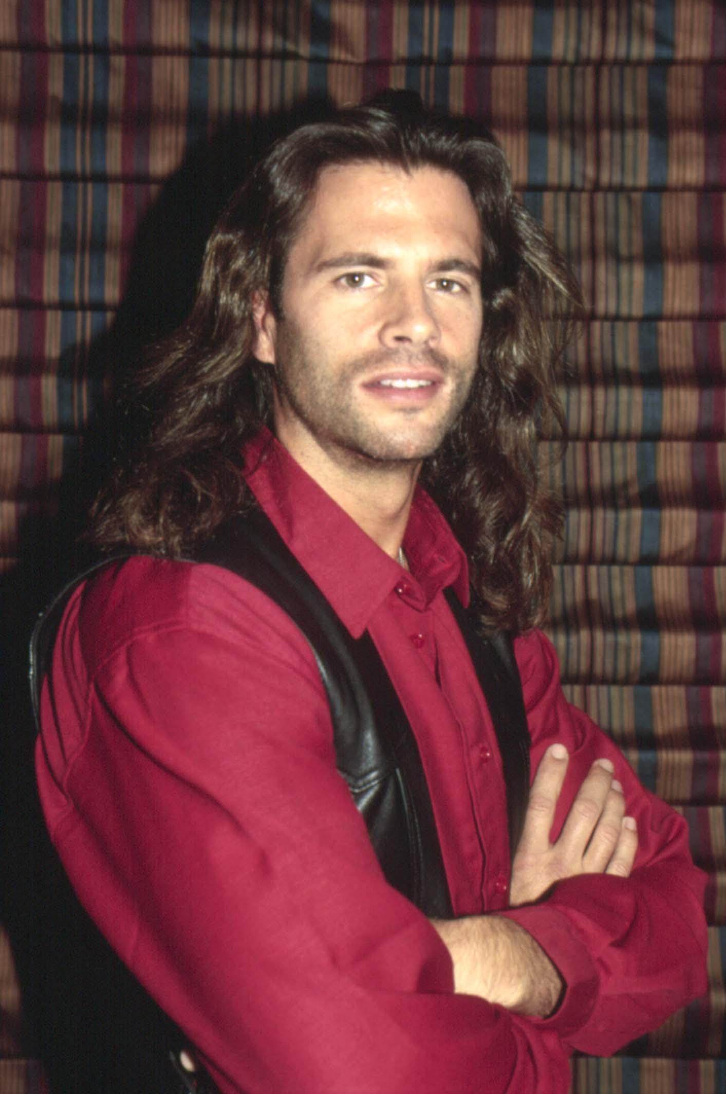 Lorenzo Lamas channels his rugged "Renegade" persona in a 1993 portrait. Posing with his arms crossed against a striped backdrop, the actor sports shoulder-length dark waves, light stubble, and his signature sharp gaze while wearing a deep red button-down shirt paired with a black leather vest. | Source: Getty Images