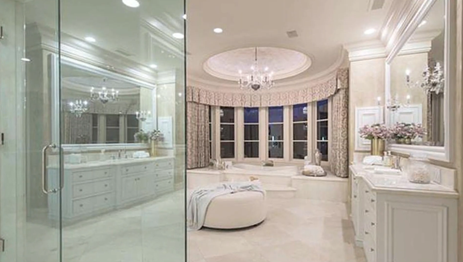 The opulent primary bathroom evokes a rotunda design with a sunken soaking tub set into a curved bay window, mirrored double vanities, an expansive glass shower, and a plush round ottoman beneath an ornate crystal chandelier. | Source: YouTube/Taste of Country
