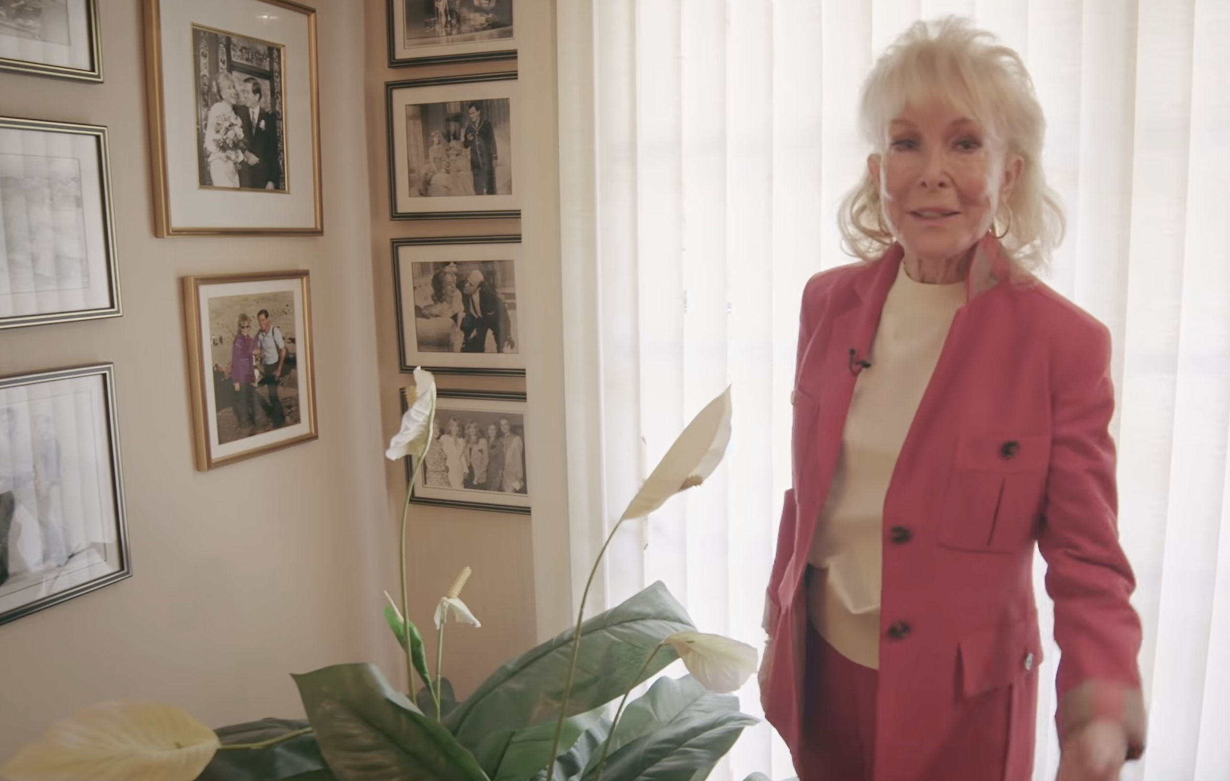 Barbara Eden poses beside a leafy houseplant in her home, wearing a pink-red pantsuit as framed photographs and career memorabilia fill the wall behind her. | Source: YouTube/People