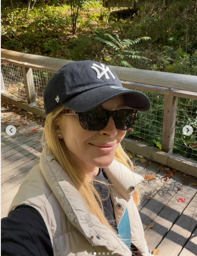 Lindsay Lohan poses in a black New York Yankees cap, oversized sunglasses, and a cream padded vest during her autumn stroll along a wooden boardwalk surrounded by greenery. | Source: Instagram/lindsaylohan