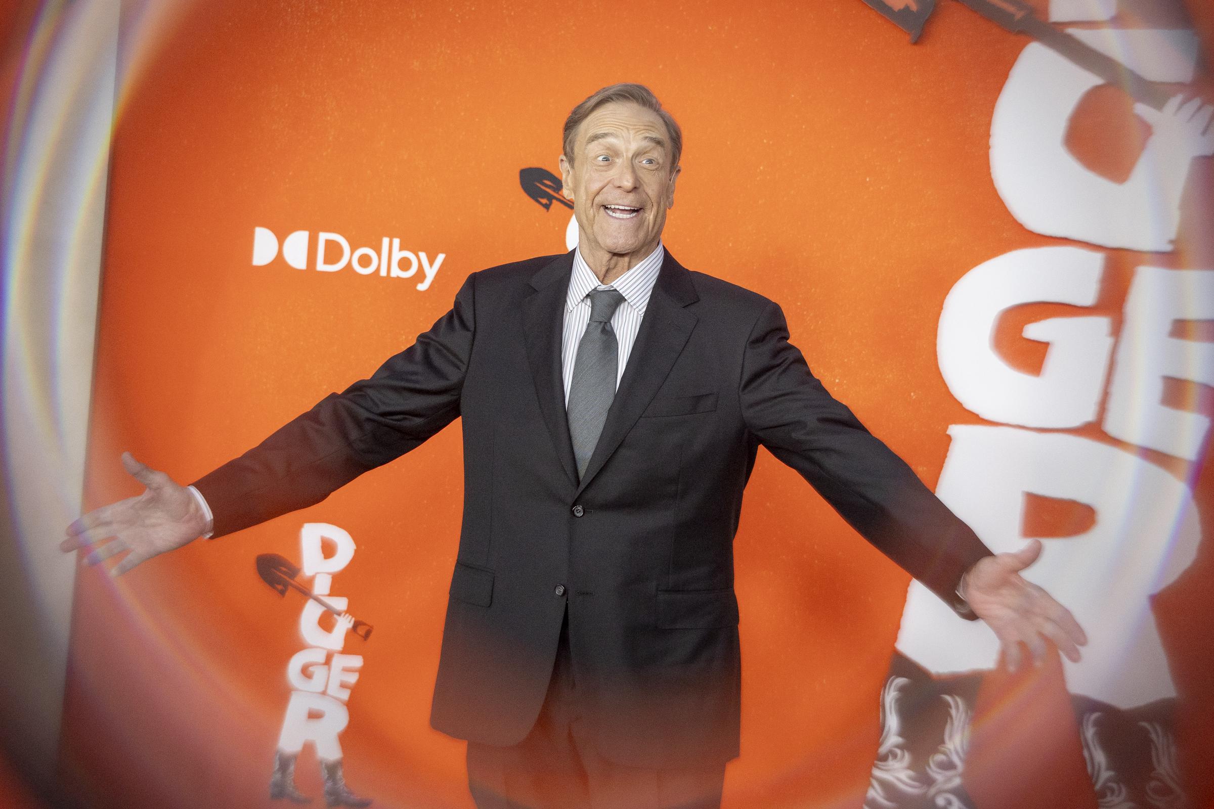 Goodman also showed a more animated side on the carpet, smiling broadly and opening his arms for photographers against the bright orange backdrop. The playful pose captures a different expression from the same premiere.