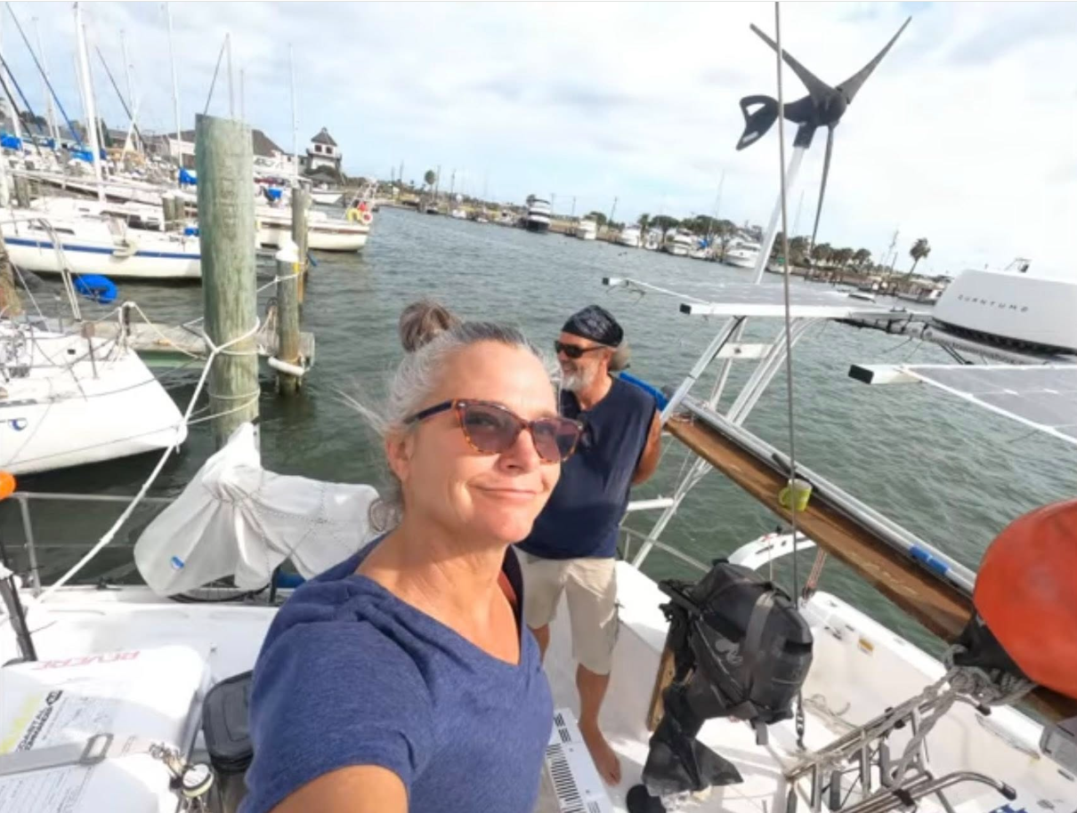Lynette Hooker takes a relaxed selfie aboard a sailboat while Brian Hooker stands behind her, both enjoying a breezy moment at a marina surrounded by docked boats and calm water. | Source: Facebook/PlunderStudios