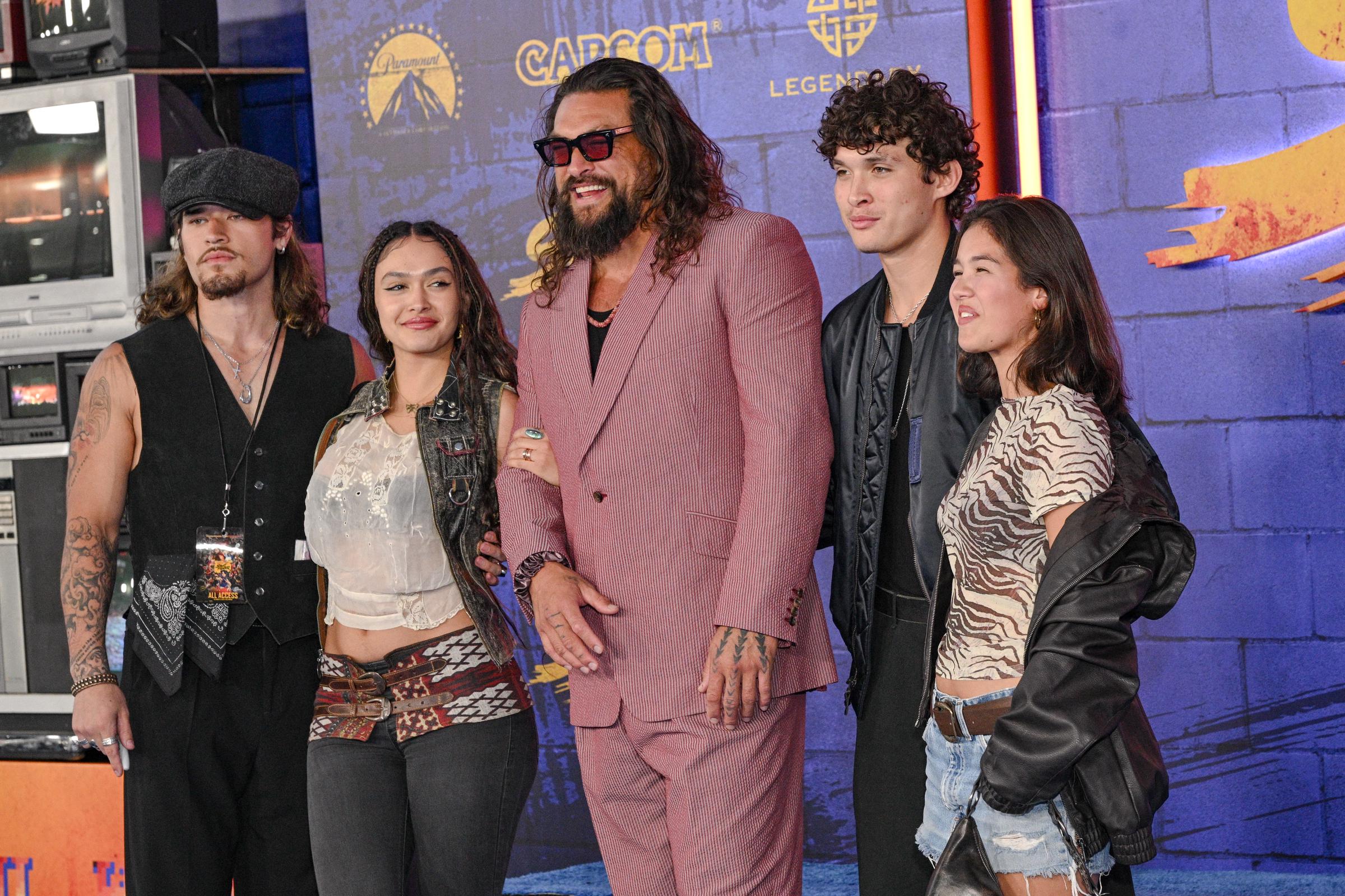Glancing off to the side toward waiting fans and press, the family shares an candid moment against the retro CRT monitors stacked beside the backdrop. The medium shot captures Momoa mid-laugh, highlighting his tinted eyewear and the textured lapels of his suit as Lola holds onto his sleeve. | Source: Getty Images