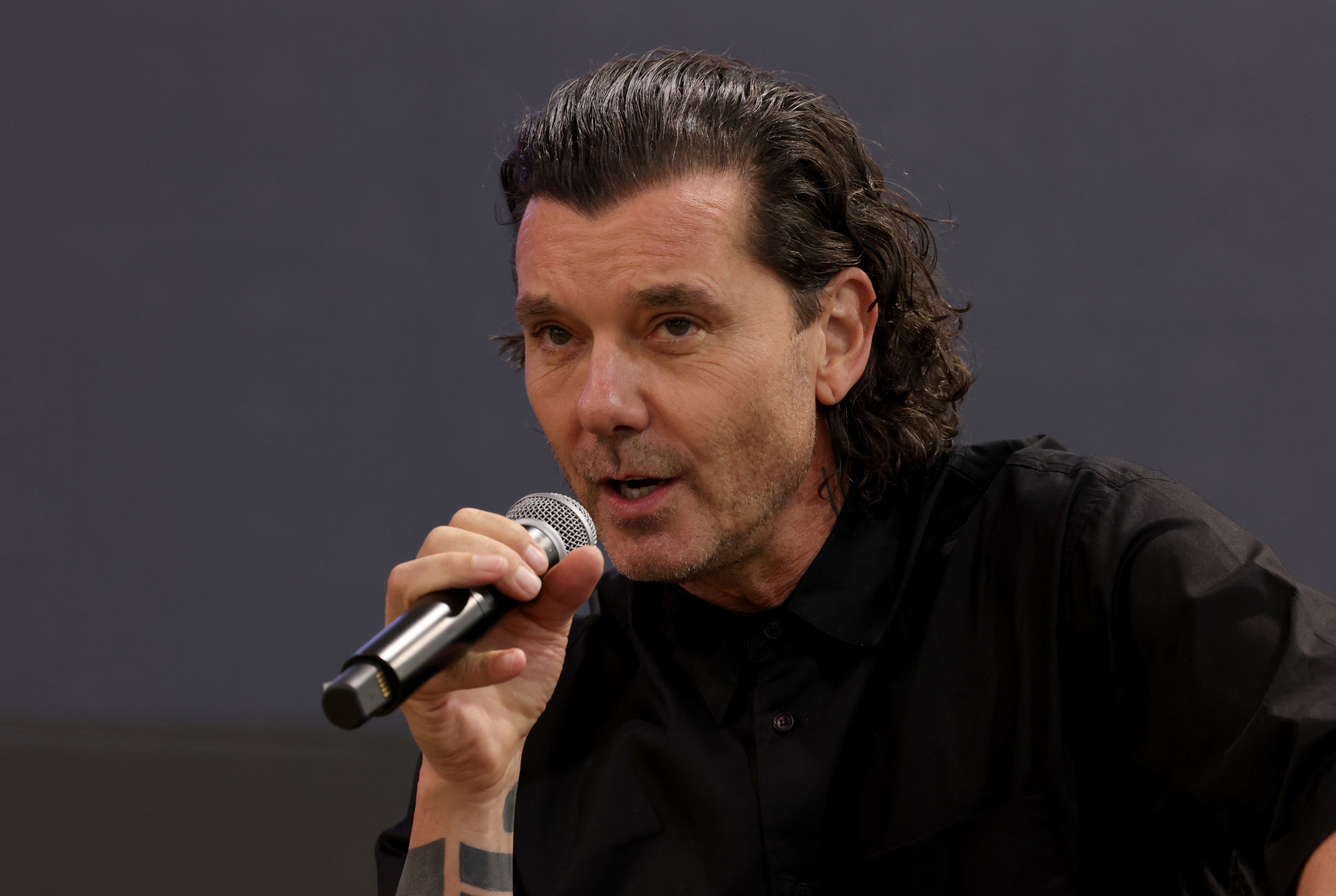 Gavin Rossdale speaks into a microphone at his "Beyond the Music" fashion debut panel in Las Vegas, wearing black as his longer hair falls back.