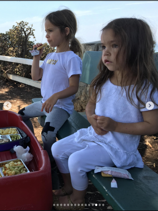 Mabel and Fiona sit together outdoors beside a red wagon filled with food during a family outing. Mabel appears to be eating while Fiona sits beside her in white, with trees and greenery surrounding them in another candid glimpse of the sisters’ childhood. | Source: Instagram/mabellowe_