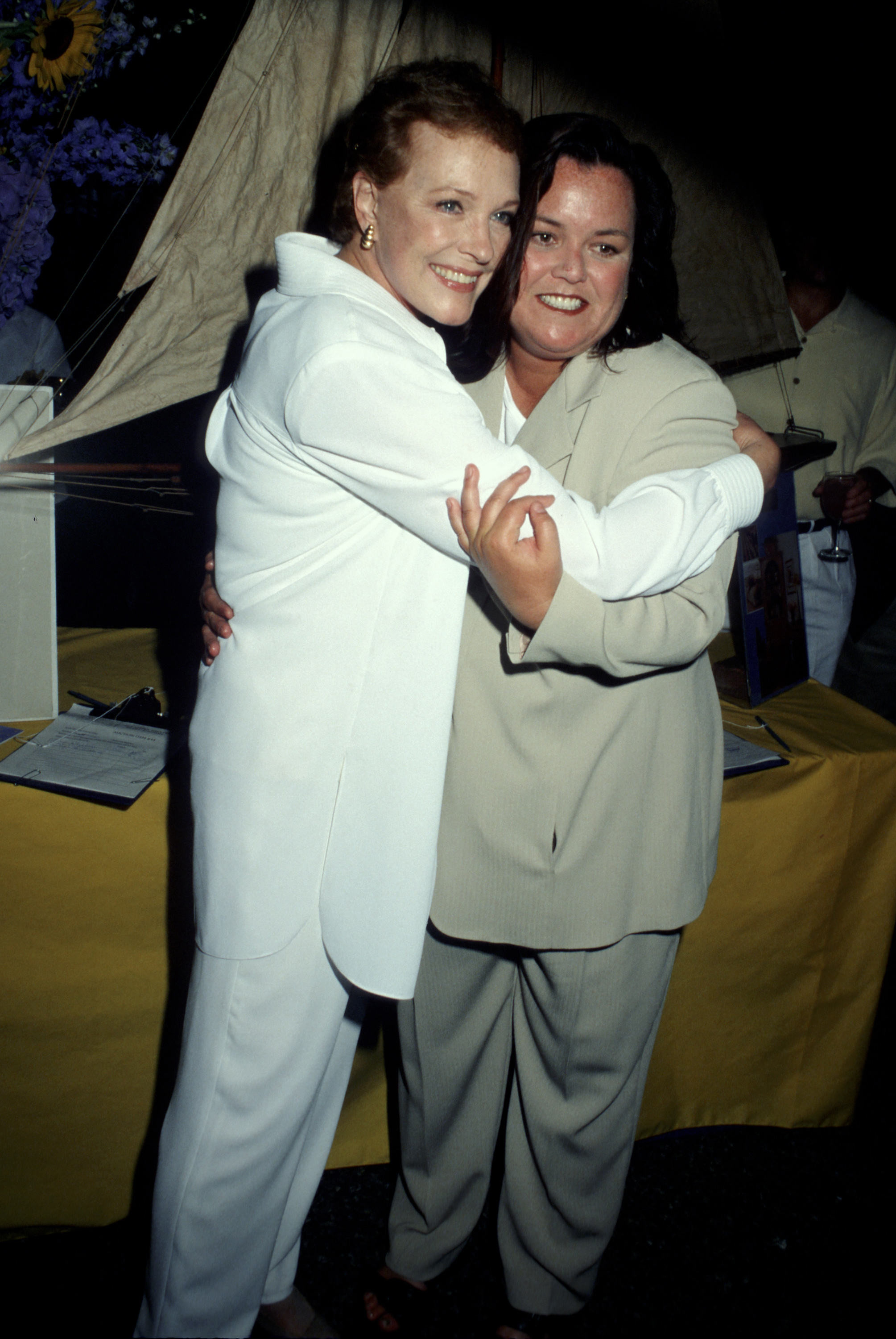 Julie Andrews shares a warm hug with comedian and television host Rosie O'Donnell during the Summer Gala Benefit Bash for the Bay Street Theatre in Sag Harbor, New York, on July 10, 1999. Andrews looks chic in an all-white ensemble featuring a collared tunic and matching trousers accented with gold hoop earrings, while O'Donnell beams in an oversized beige pant suit.