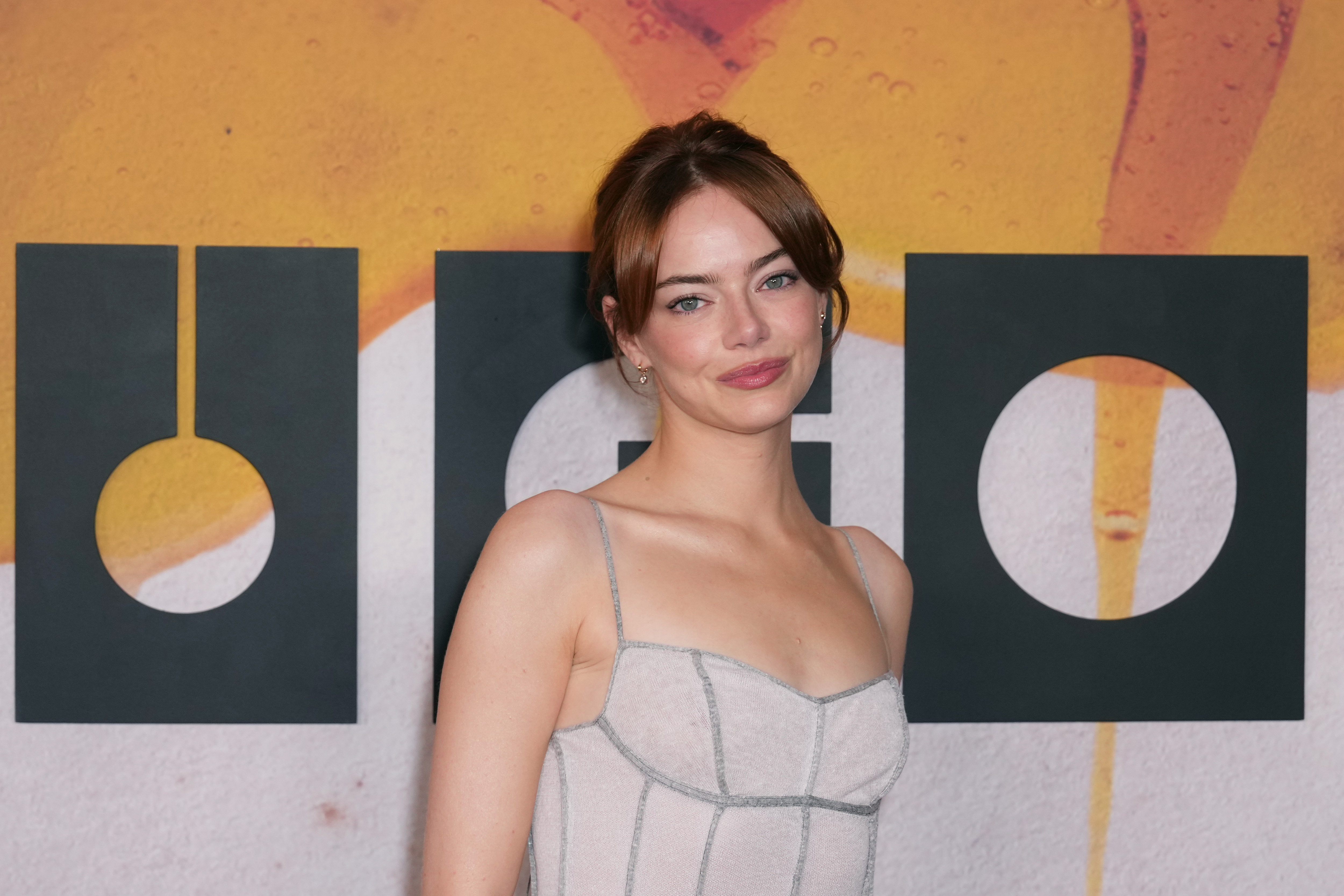 Emma Stone poses with confidence, her shoulders angled and lips curved in a knowing half-smile. The soft sheen of her gown catches the light against the bold "Bugonia" backdrop.