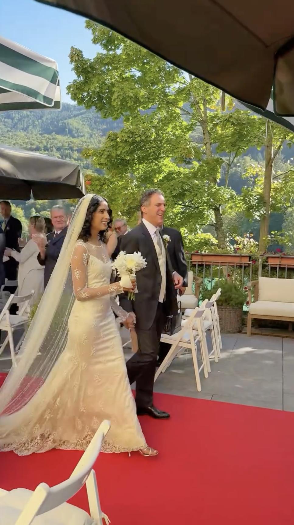 Sheela and Alexander Newley walk the aisle, greeted by applause. Sheela wears a lace gown and veil while carrying a white bouquet, beaming beside her husband. | Source: Instagram/sheelanewley