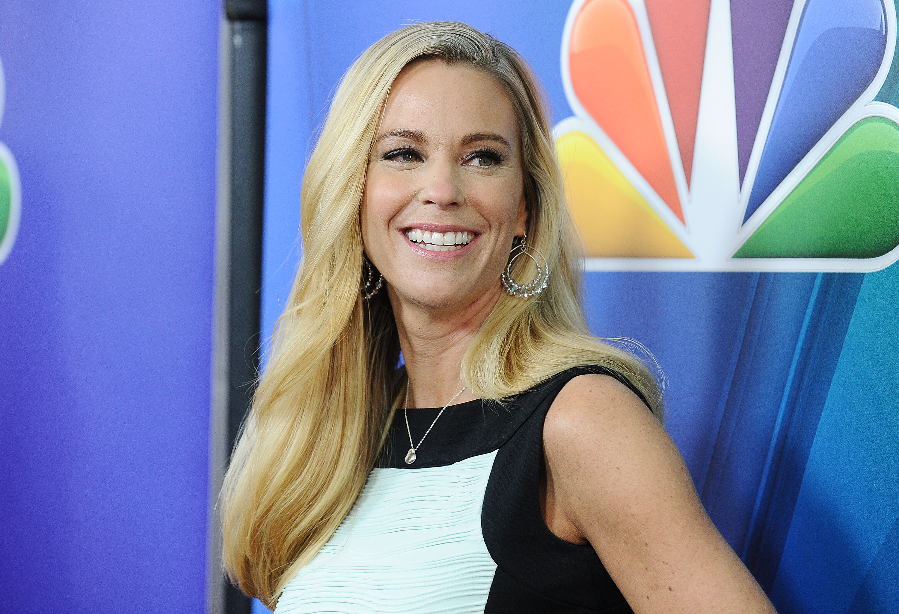 At an NBCUniversal press event, Kate Gosselin glows in a sleeveless black-and-white dress. With flowing blonde hair, hoop earrings, and a radiant smile, she steps out with a polished look for the cameras.