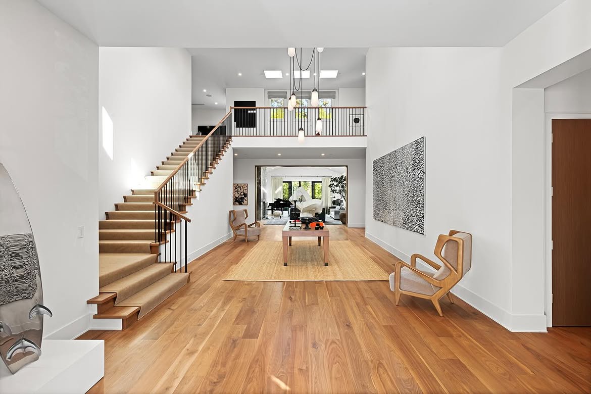 The later entry keeps the soaring proportions of the earlier foyer but introduces a different mix of furnishings and artwork. A runner now follows the staircase, while sculptural chairs and a large monochrome piece give the open space a more collected appearance. | Source: Instagram/theshevins
