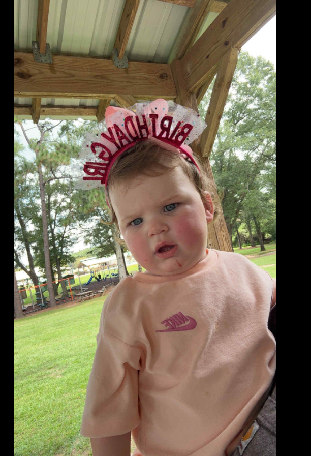 Keziah Luker's 18-month-old toddler stands outdoors under a wooden shelter, wearing a soft peach outfit and a sparkly headband. | Source: Facebook/Keziah Luker
