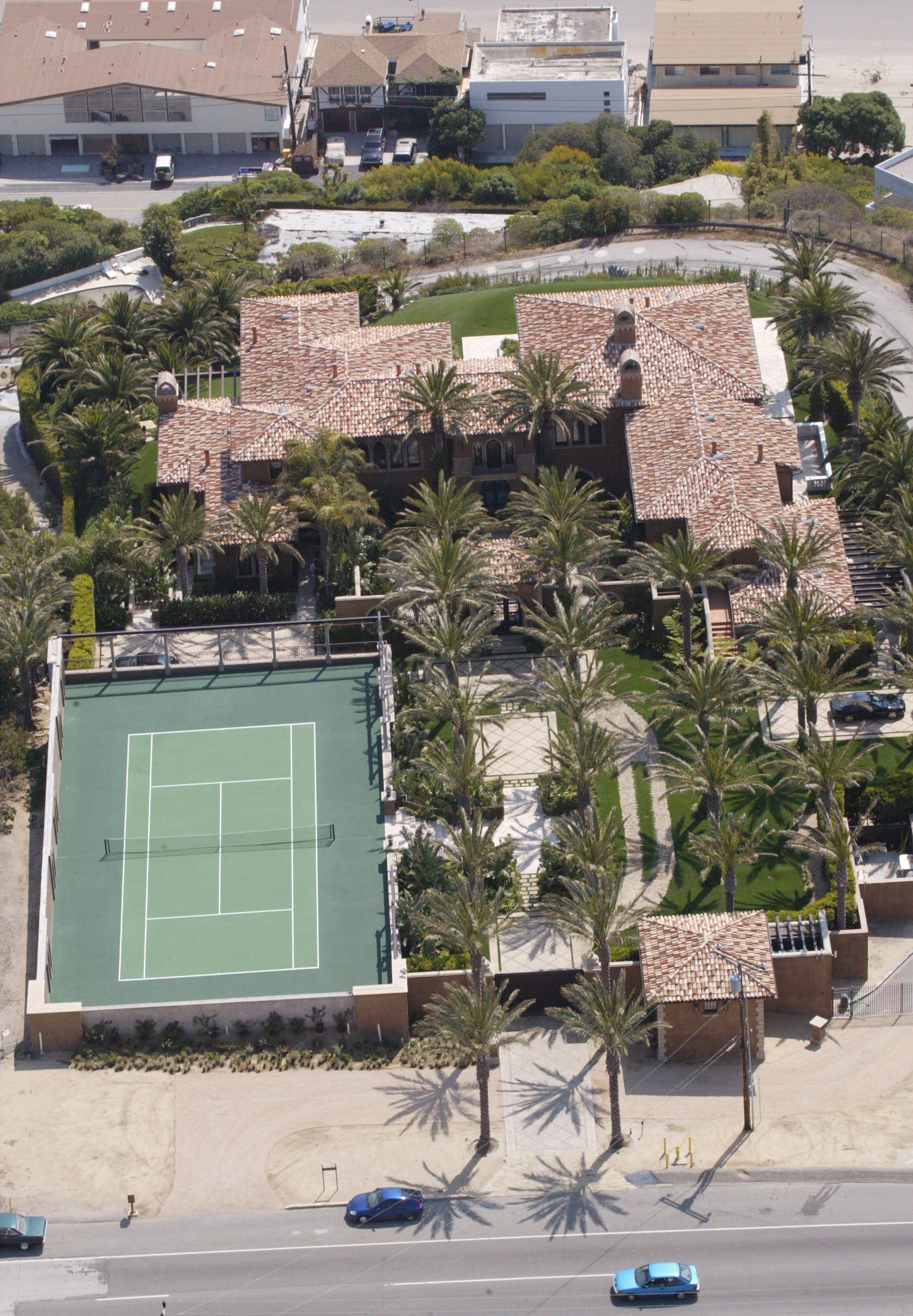 An overhead aerial view captured actress Cher's gated estate in Malibu, California, on March 27, 2002. The property features extensive terracotta-tiled roofs, a private green tennis court, and manicured walkways lined with towering palm trees positioned alongside the highway. | Source: Getty Images