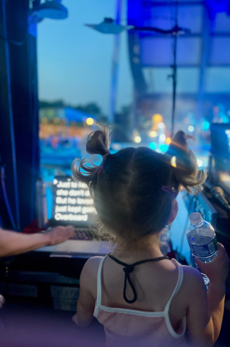 Kaliyah stands near the side of a stage, holding a water bottle as blue lights illuminate the performance area beyond her. A laptop displaying performance information sits nearby, showing the behind-the-scenes environment she experiences while traveling with Wynonna. | Source: Instagram/wynonnajudd